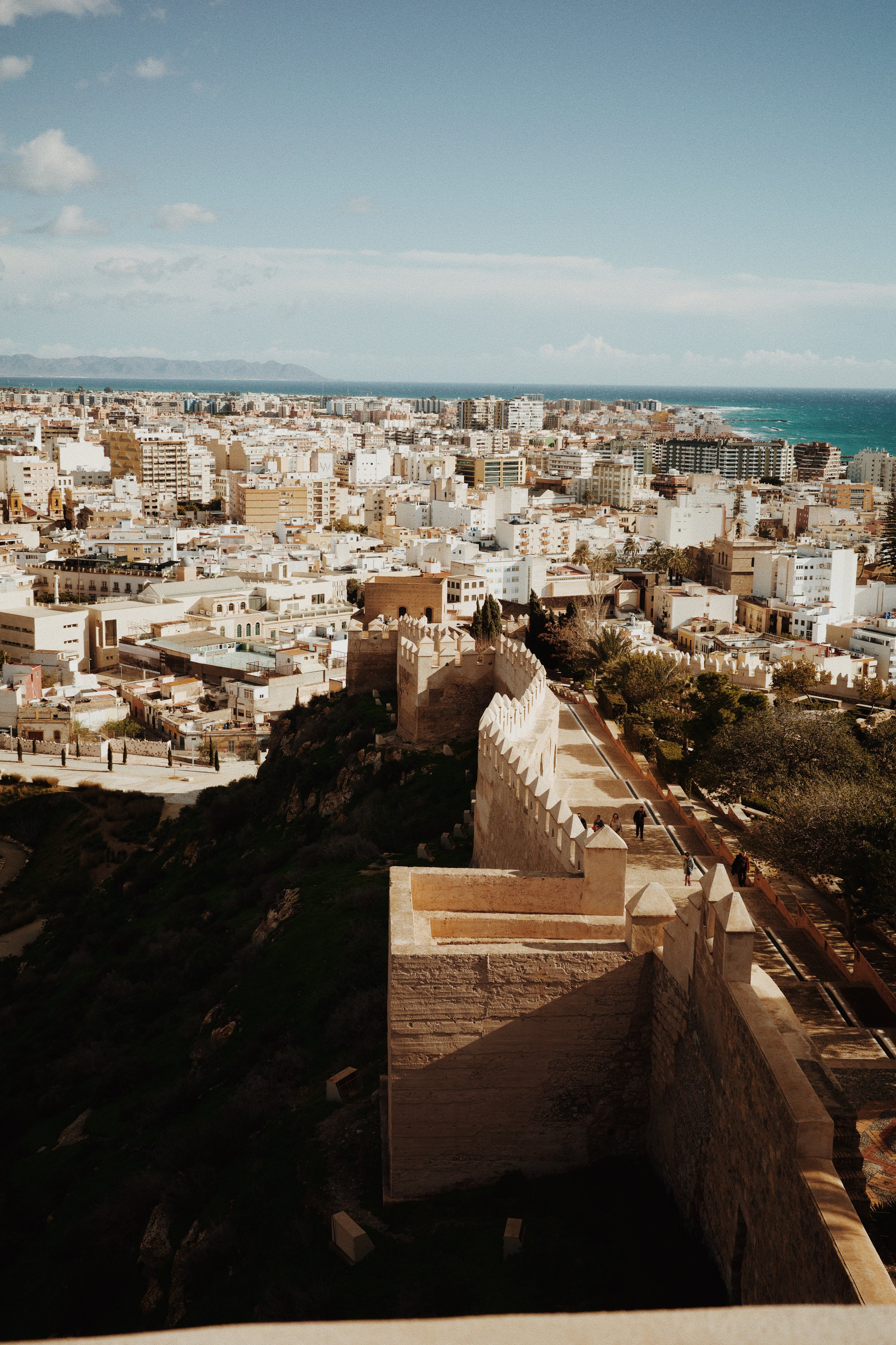 Vue aérienne d'une ville côtière avec des bâtiments blancs et un château, surplombant la mer turquoise et un ciel bleu.