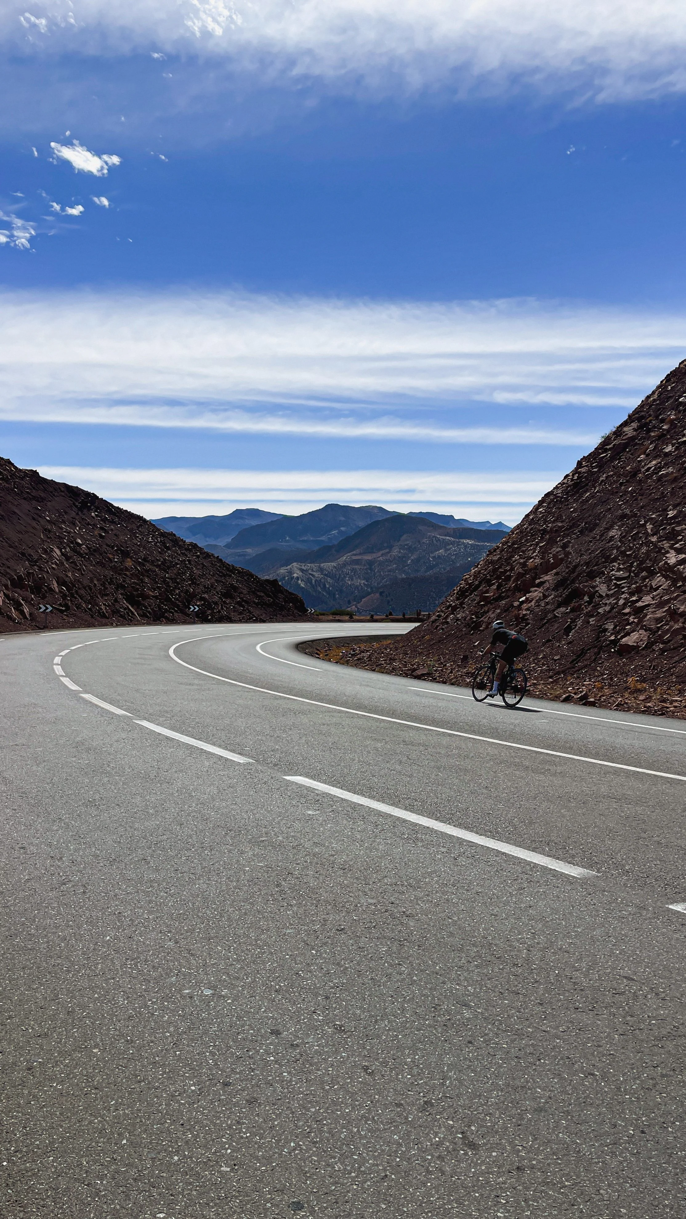 Un cycliste roule sur une route sinueuse entourée de montagnes rocheuses sous un ciel partiellement nuageux.