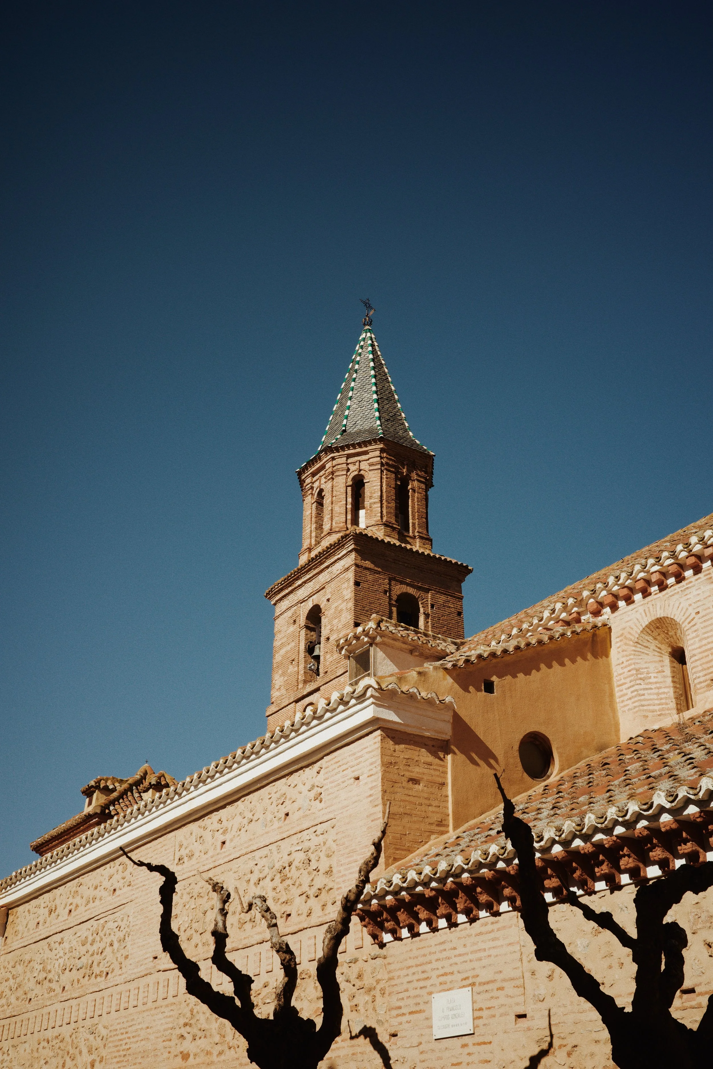 Église en pierre avec un clocher pointu, sous un ciel bleu clair, avec des arbres taillés en forme d'arc au premier plan.