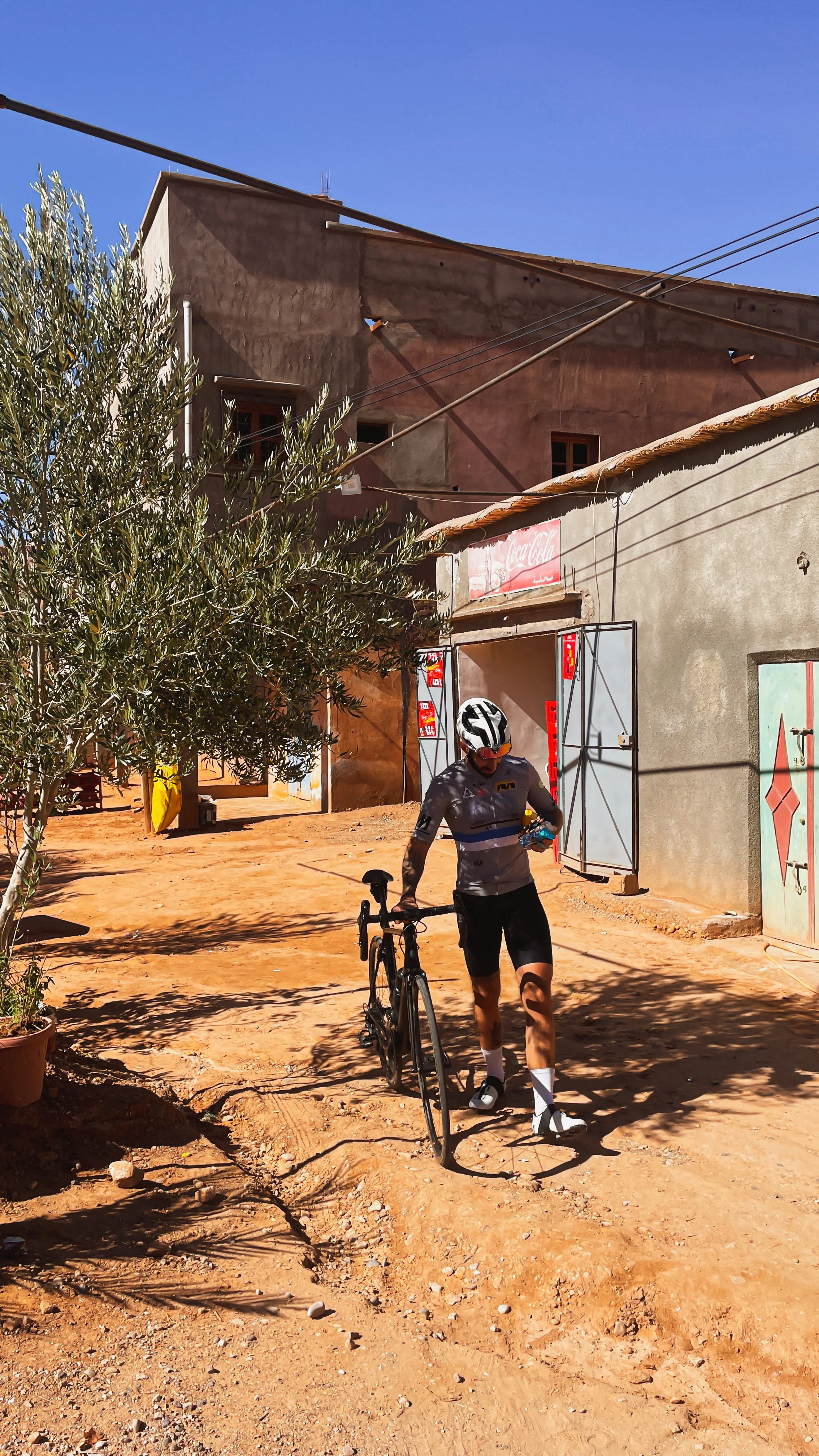 Un cycliste portant un casque, un maillot gris, des pantalons courts noirs et des chaussures de cyclisme, marche avec son vélo dans une rue en terre rouge, sous un ciel clair. En arrière-plan, il y a des bâtiments en pierre ou en ciment, une porte en fer, une feuille d'arbre vert et un panneau Coca-Cola.