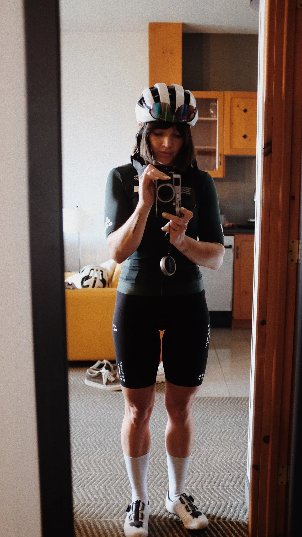 Jeune femme en tenue de cyclisme avec casque, lunettes de soleil sur la casque, portant un maillot noir, shorts de cyclisme noirs, chaussettes blanches et chaussures de cyclisme, se prenant en selfie devant un miroir dans un hôtel.