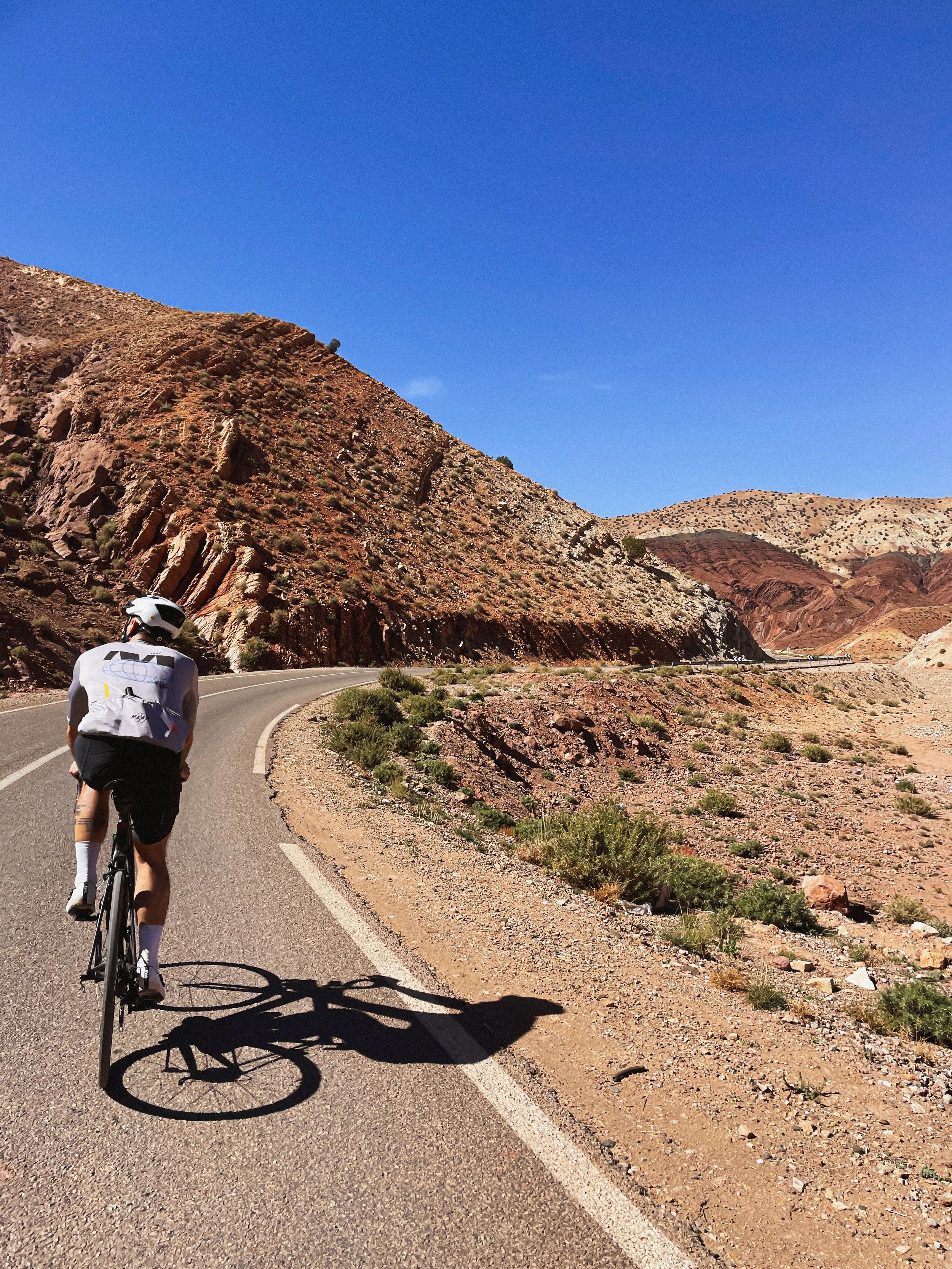 Coureur cycliste sur une route sinueuse dans un paysage désertique avec des collines rocheuses et un ciel bleu clair.