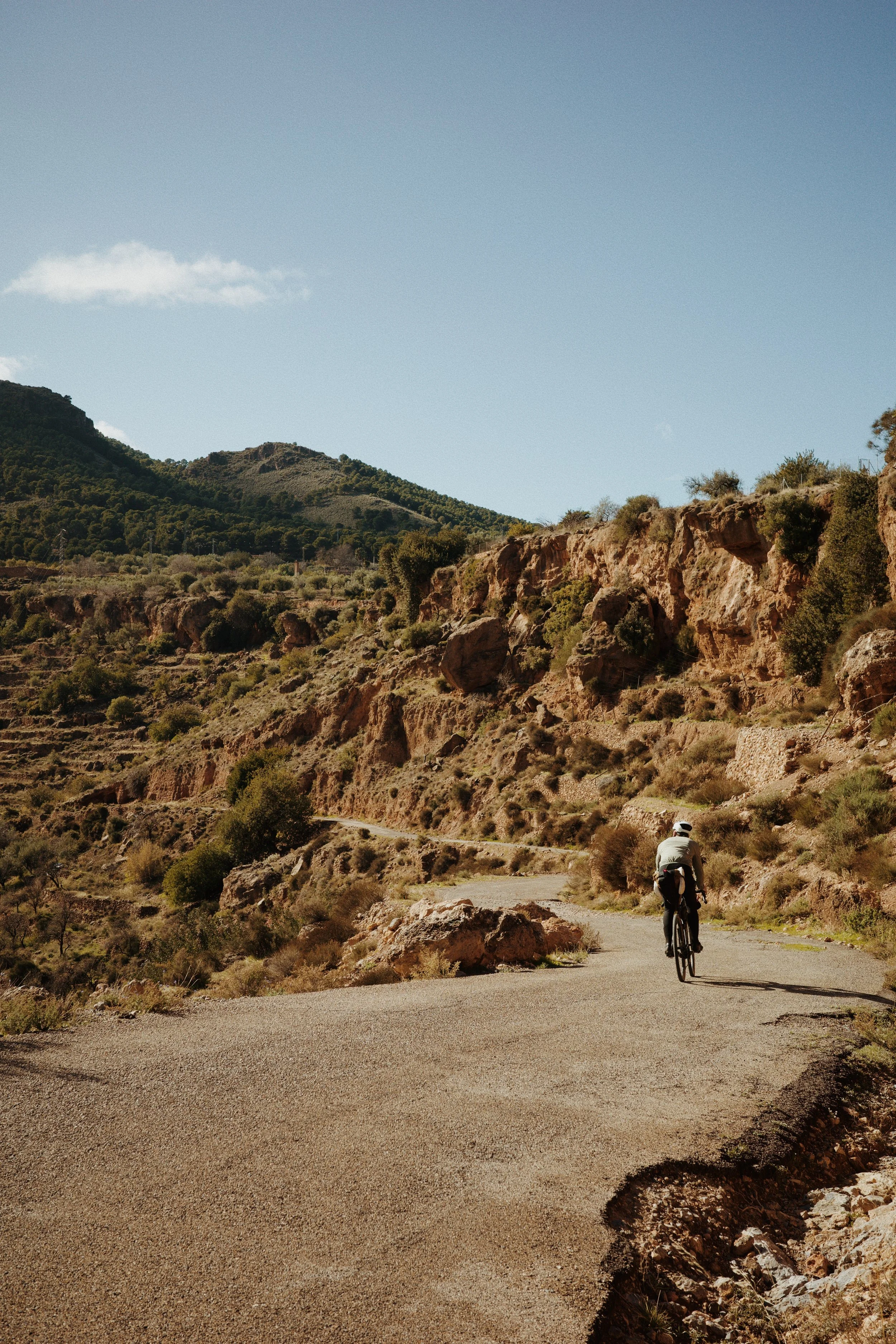 Coureur de vélo pédalant sur une route de montagne en terrain accidenté avec des collines et des rochers, sous un ciel bleu clair.