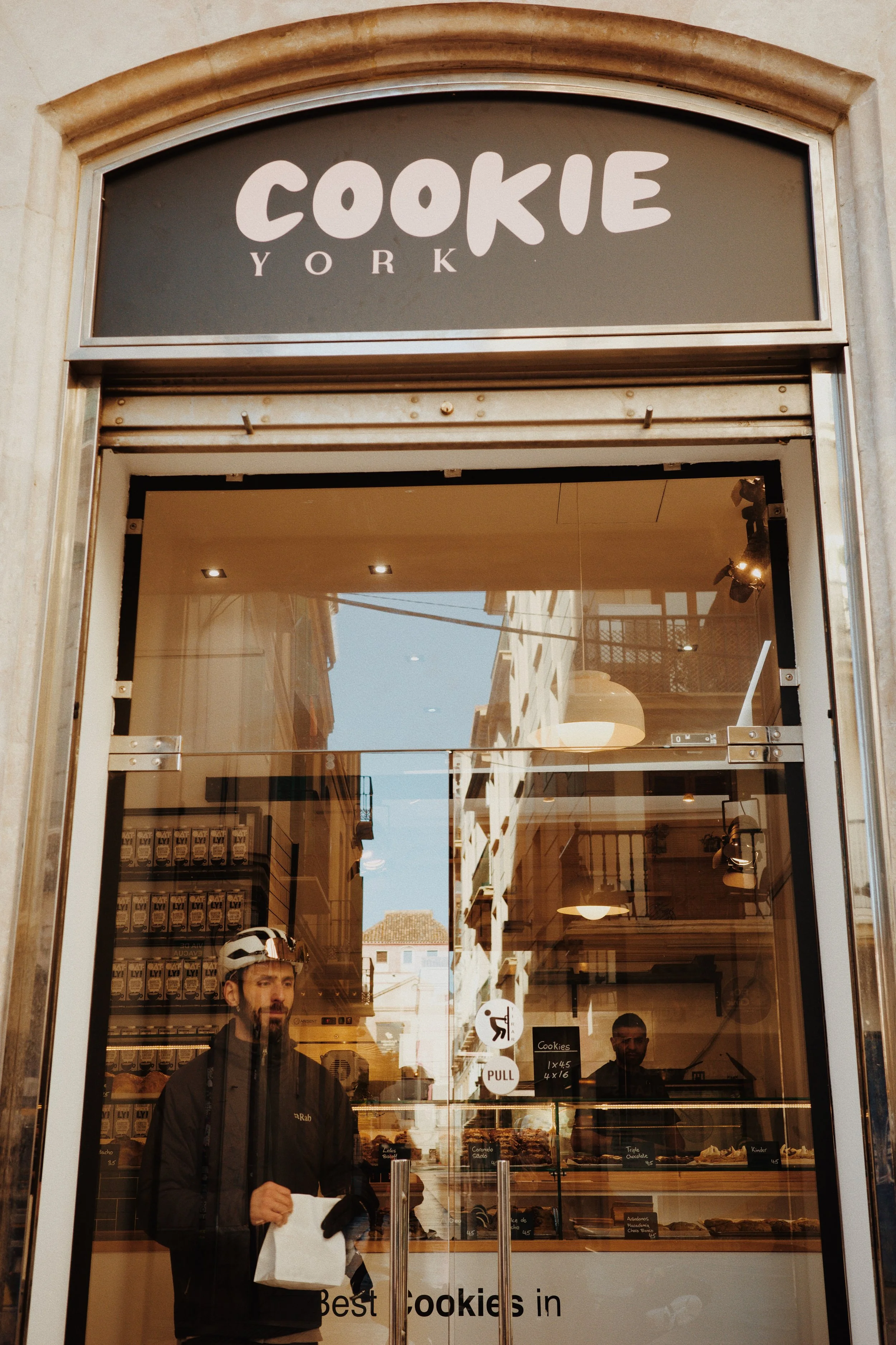 Vue de l'extérieur d'une boutique de cookies appelée 'COOKIE YORK'. À l'intérieur, deux personnes sont visibles, une portant un casque de vélo et une veste noire, tenant un sac blanc. La vitrine montre plusieurs types de cookies exposés.