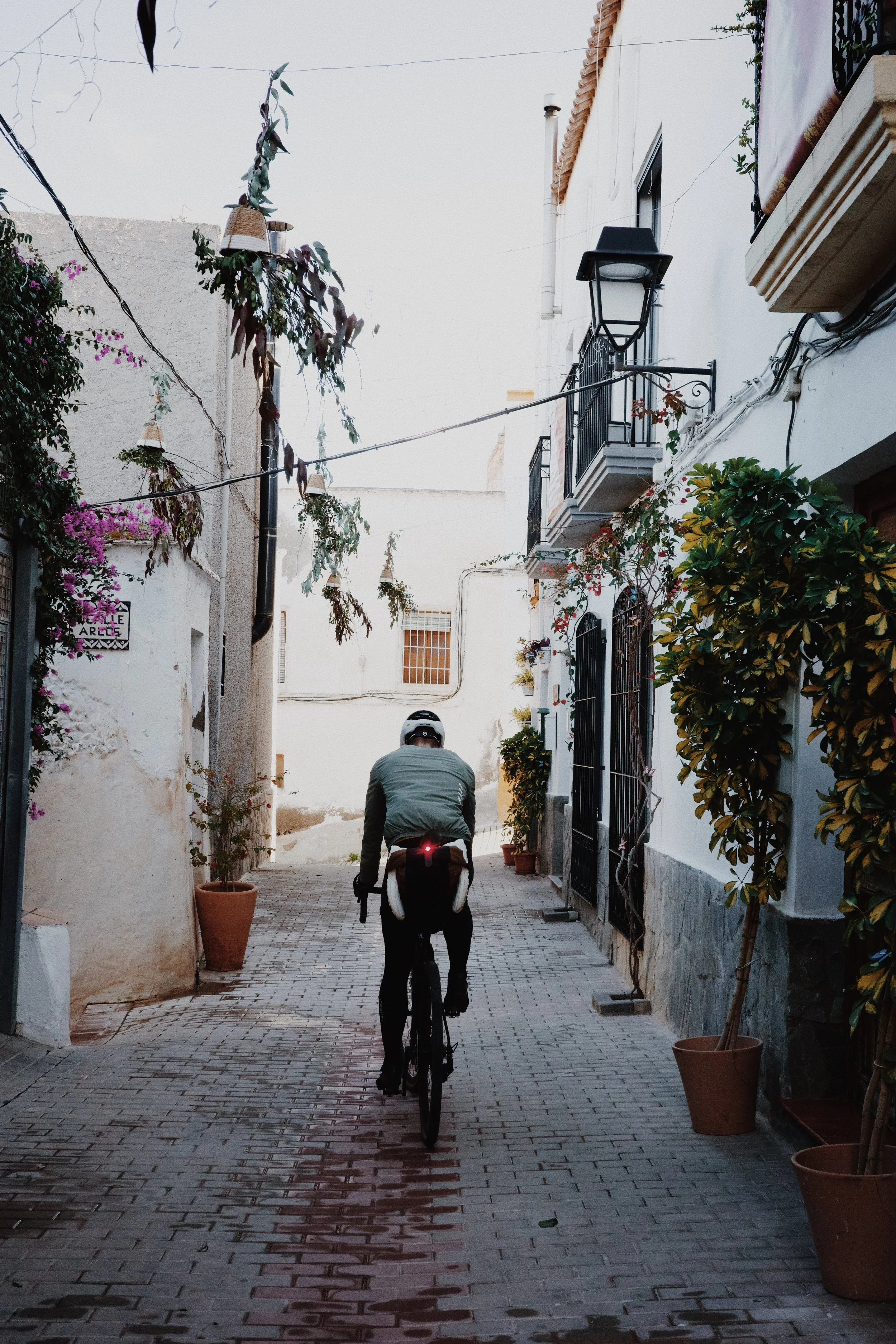 Un cycliste portant un casque traverse une ruelle pavée bordée de maisons blanches et de plantes en pot, avec un ciel clair en arrière-plan.