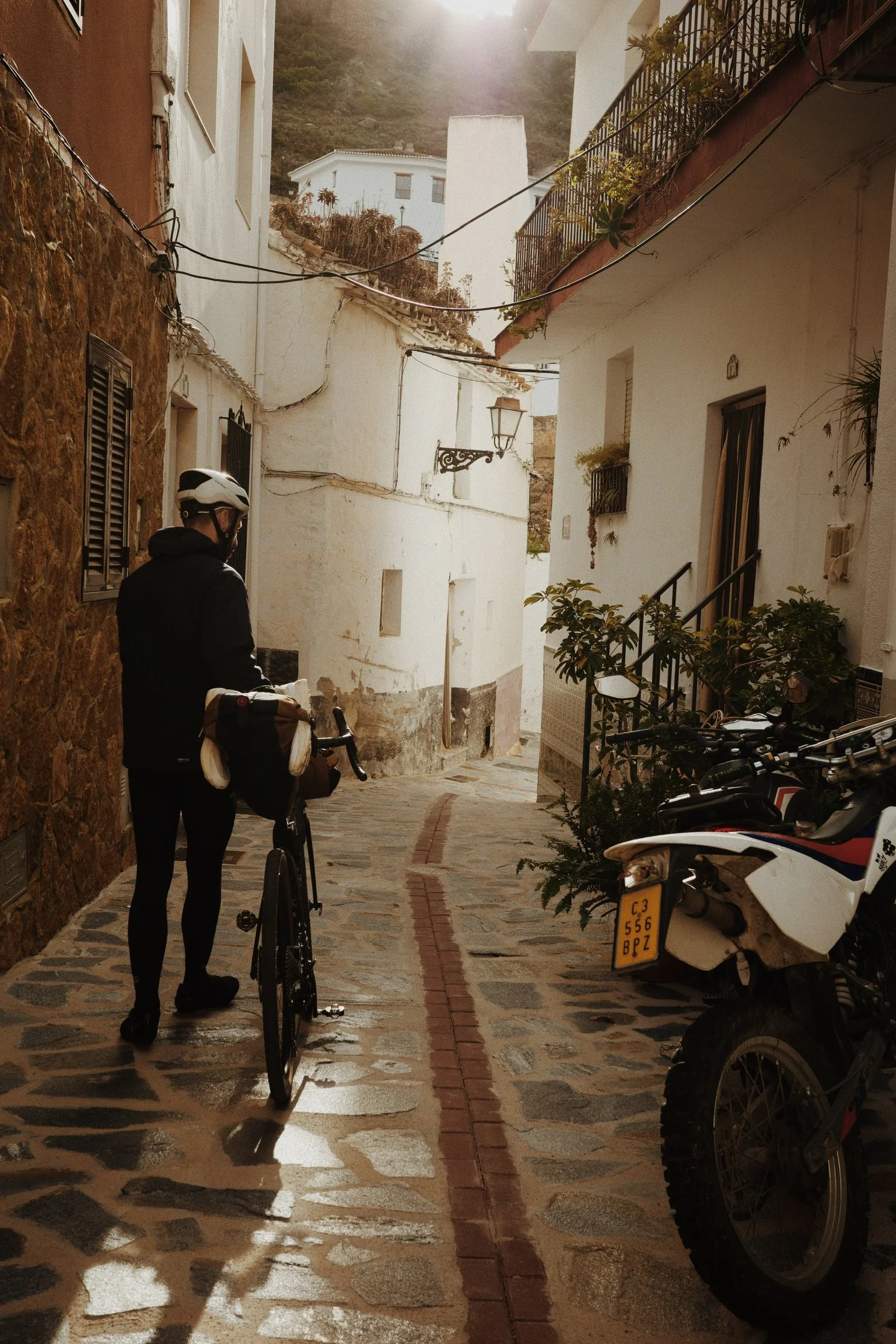 Une rue pavée étroite avec un homme portant un casque et des vêtements noirs, se tenant près d’un vélo. À droite, plusieurs motos garées, et à gauche, un mur en pierre. Sur la gauche, une maison blanche avec des fenêtres à volets noirs, et à droite, une maison blanche avec un balcon en fer forgé. Le ciel est en partie visible avec la lumière du soleil.