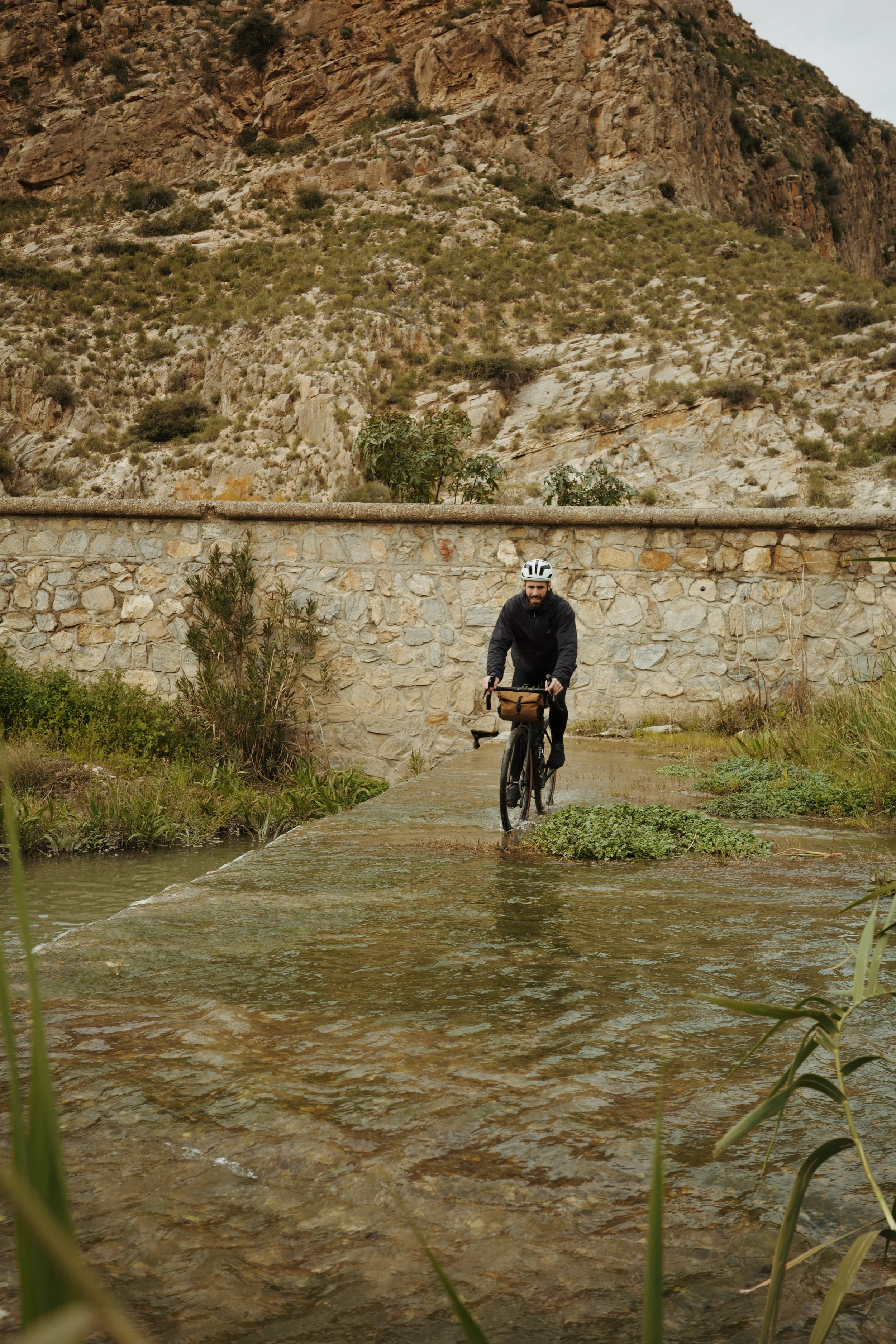 Un homme en casque fait du vélo sur un chemin en pavé, traversant une rivière peu profonde avec des montagnes rocheuses en arrière-plan.