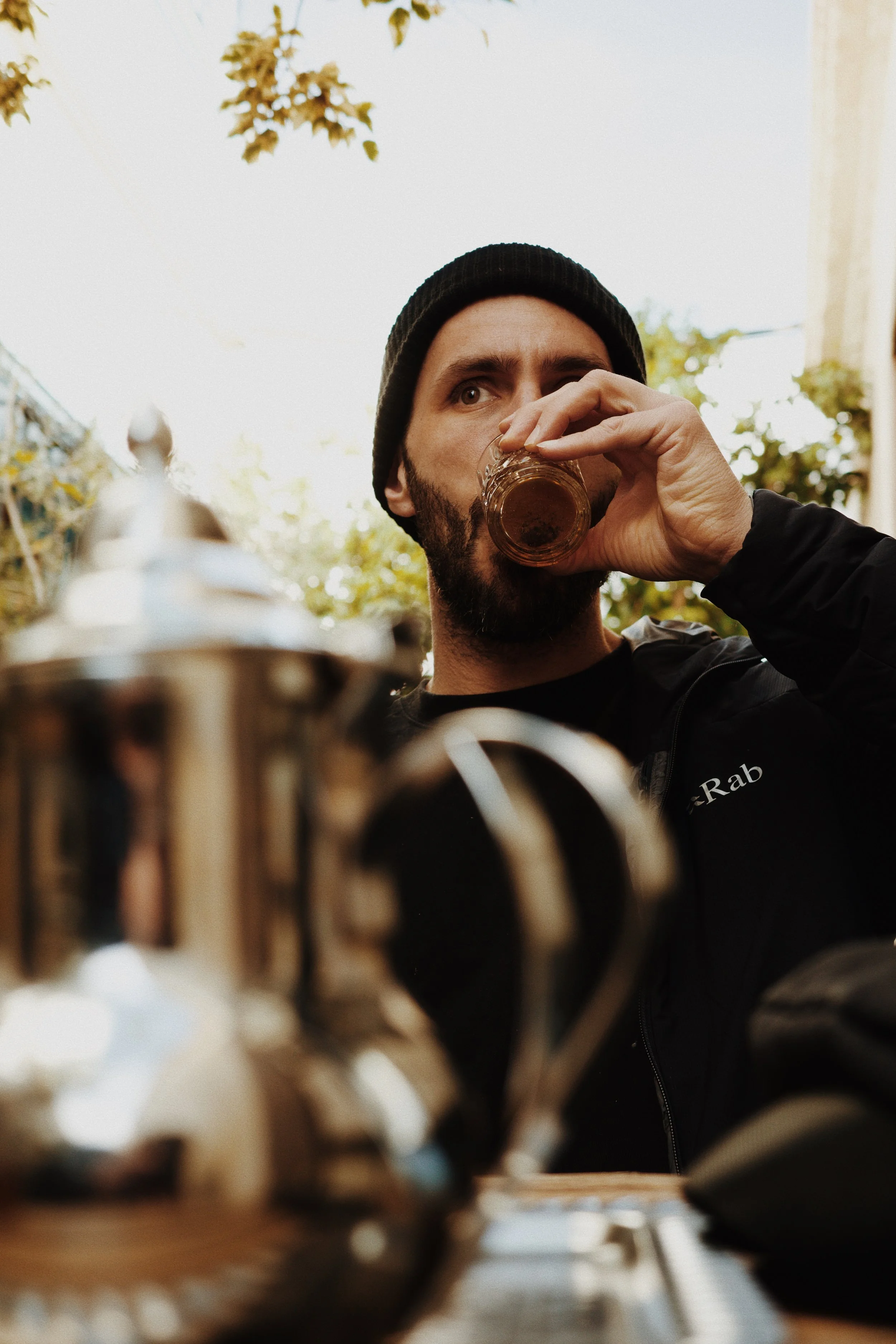 Jeune homme avec un bonnet noir buvant une boisson dans un verre en verre, assis à une table avec des objets en laiton ou en métal en premier plan, à l'extérieur avec ciel clair et feuillage en arrière-plan.