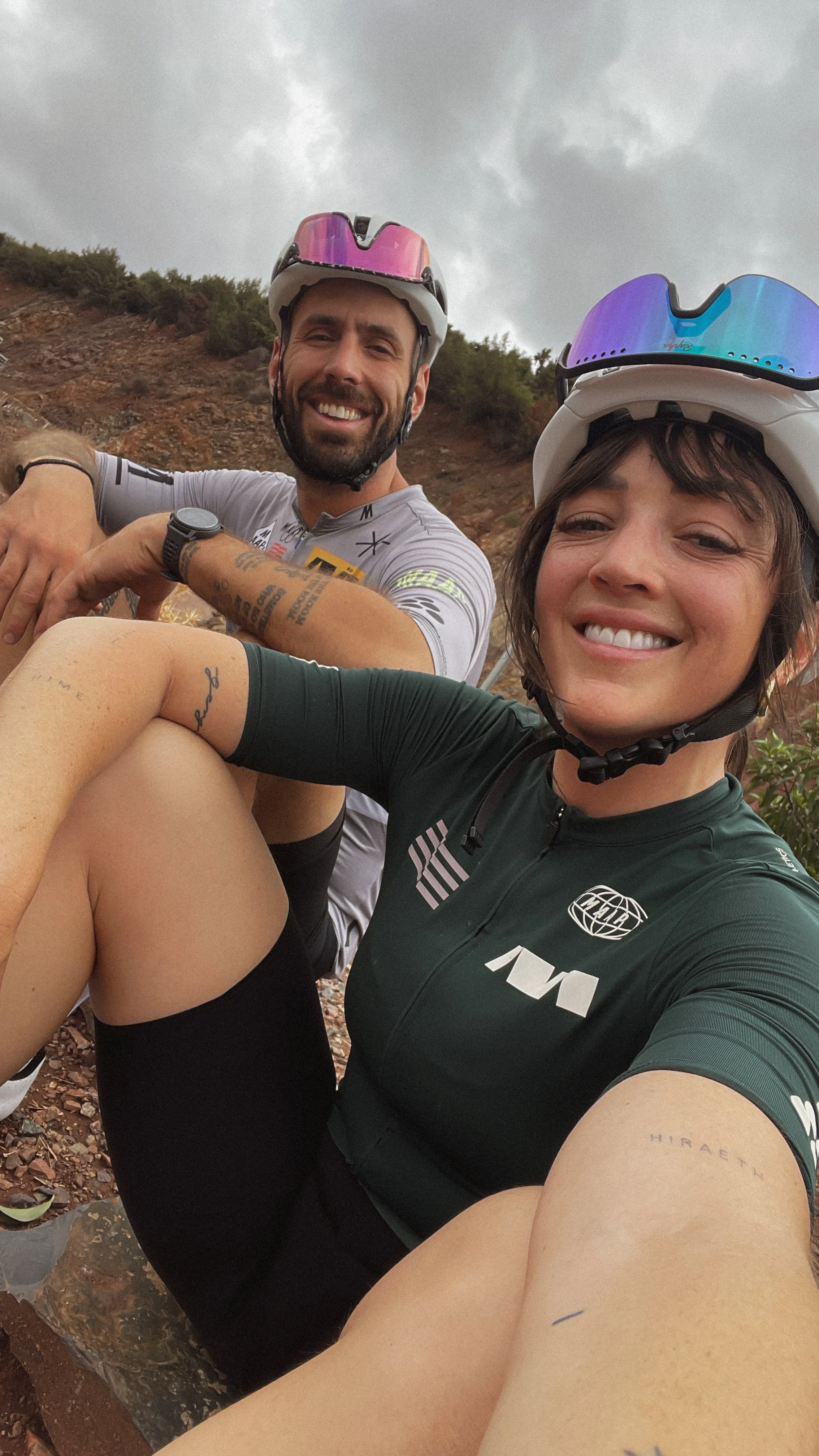 Deux cyclistes portant des casques et vétements de cyclisme prennent un selfie lors d'une sortie en plein air avec un ciel nuageux en arrière-plan.