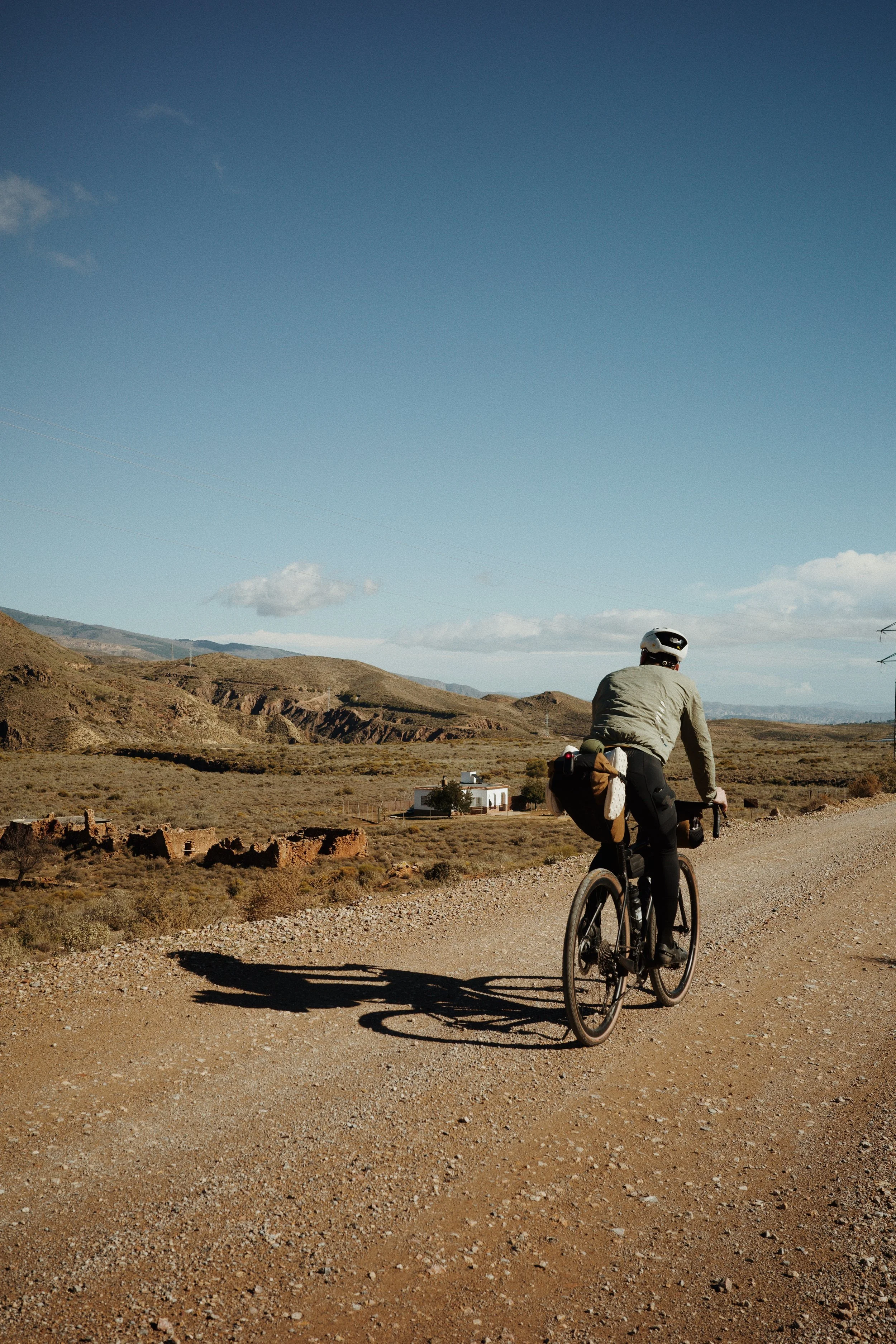 Un cycliste portant un casque, vu de dos, pédale sur un chemin de terre dans un paysage désertique avec des collines et un ciel bleu clair.