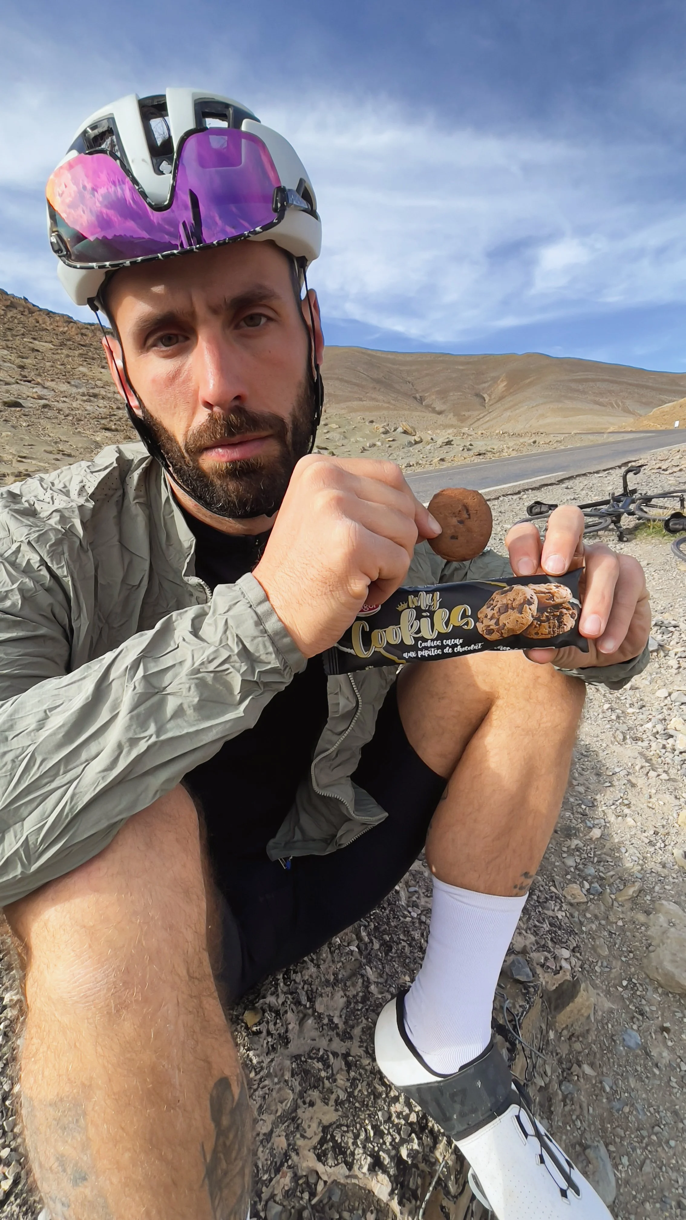 Un homme en tenue de cyclisme avec un casque, assis sur un sol rocheux dans un paysage désertique, tenant un cookie chocolaté dans une main et une barre de cookies dans l'autre.