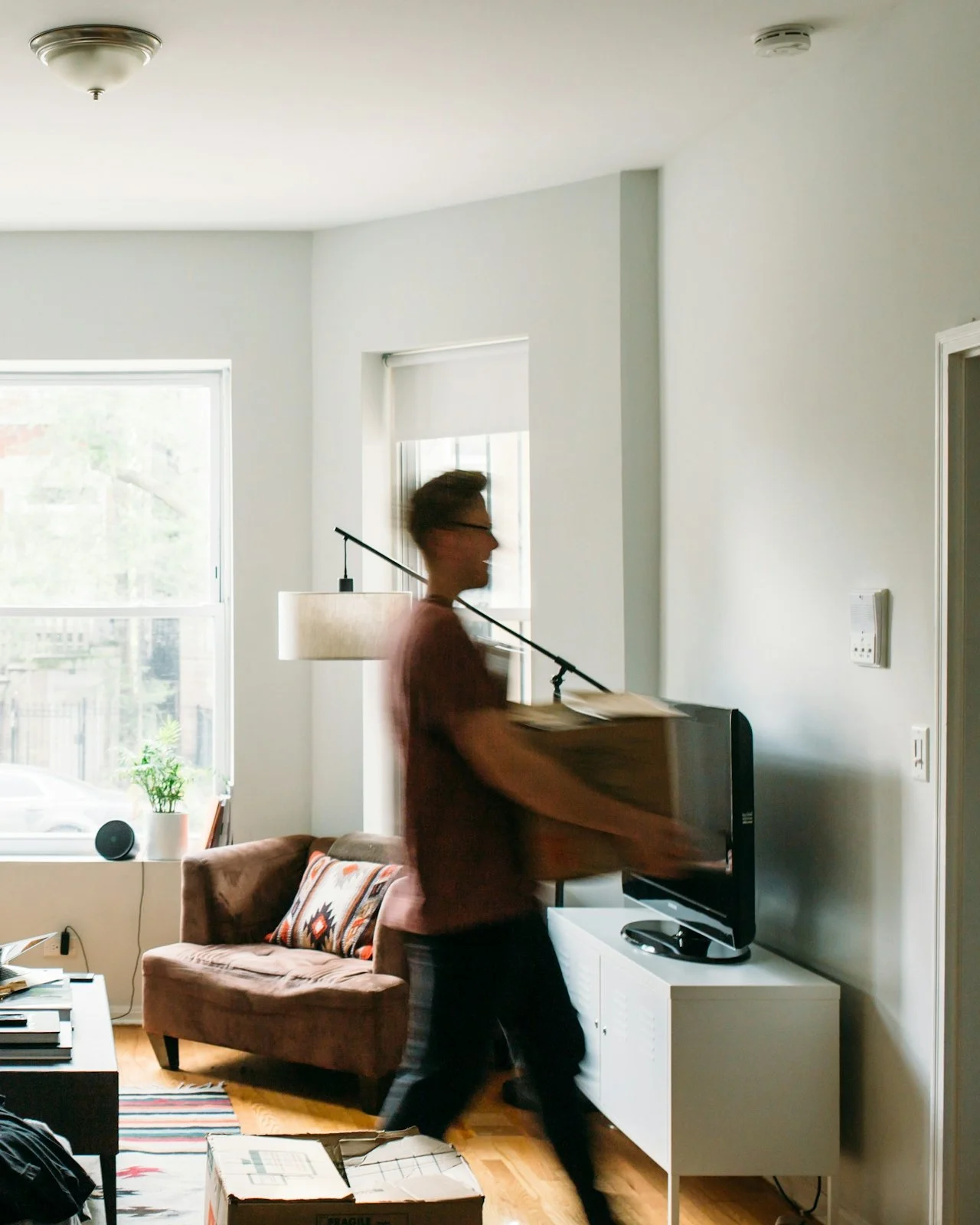 A person carrying a box across a living room with a brown couch, decorative pillows, a window, a TV on a white stand, and a potted plant.
