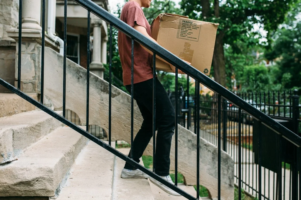 Person carrying a cardboard box walking up outdoor stone stairs with a black metal railing.