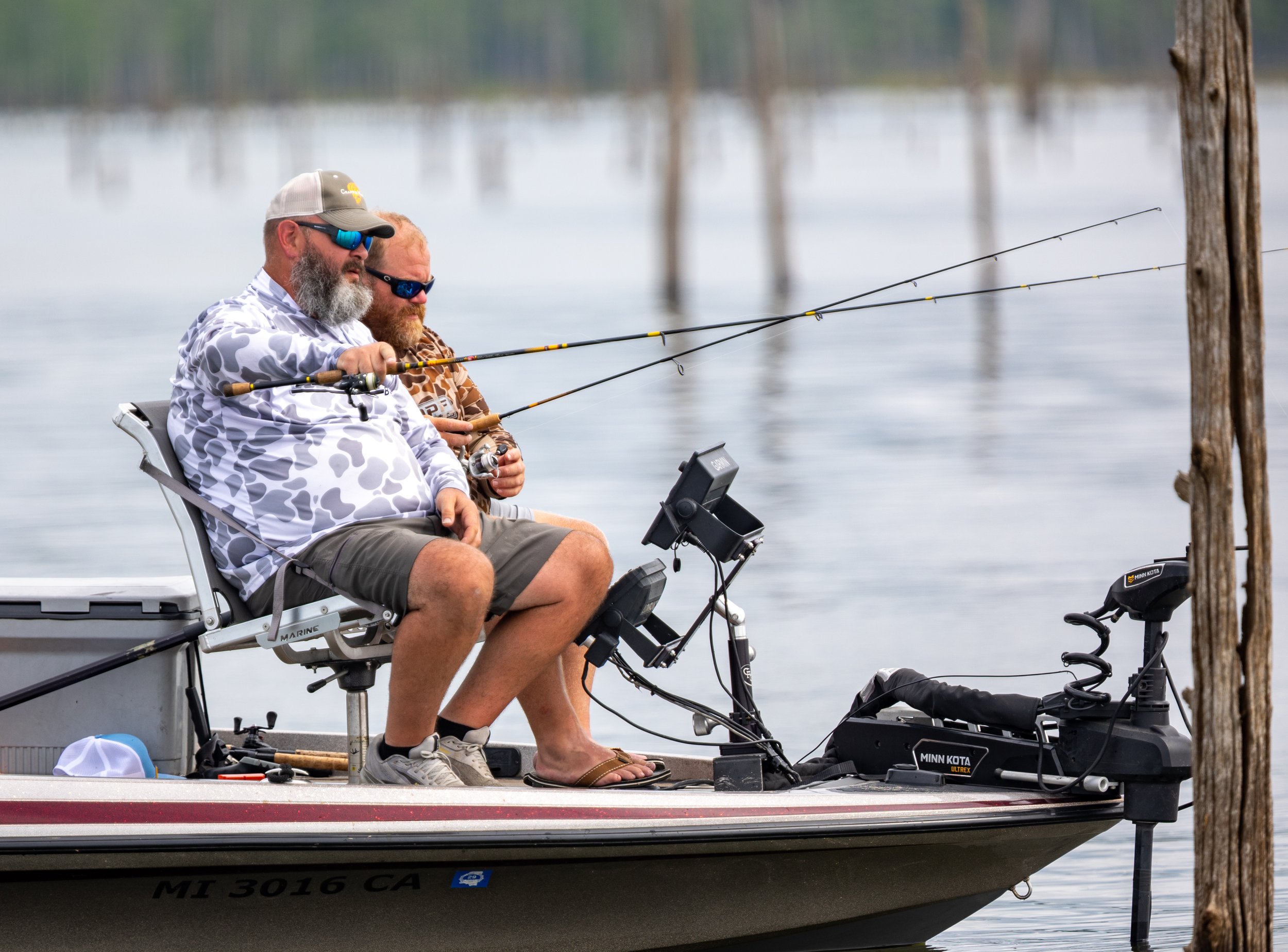 Two men sitting in a boat fishing by a lake, with fishing rods extended over the water and wooden posts in the background.