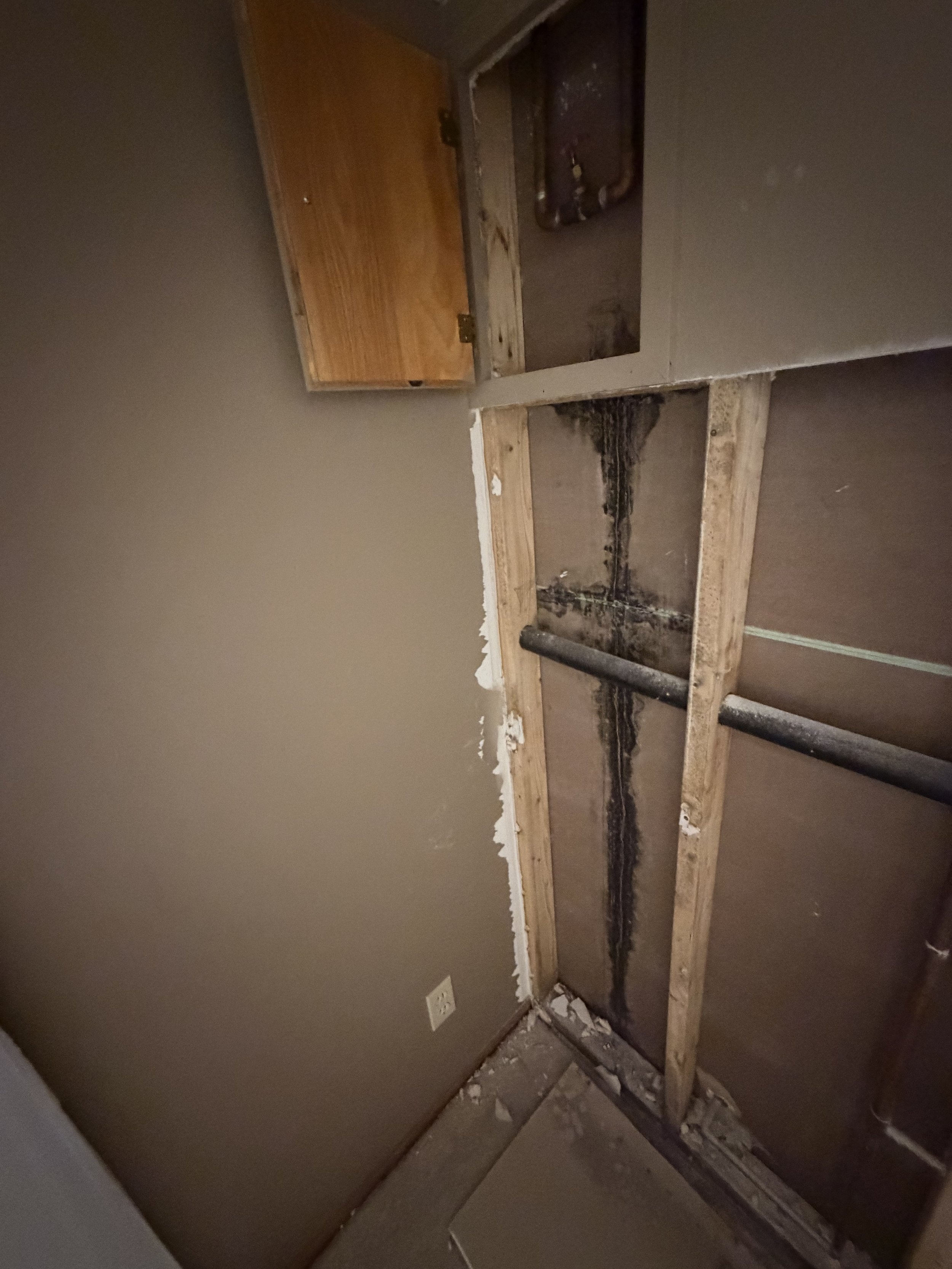 A partially demolished bathroom area, showing exposed framing and plumbing with a toilet drain pipe, and a wooden cabinet with a closed door.