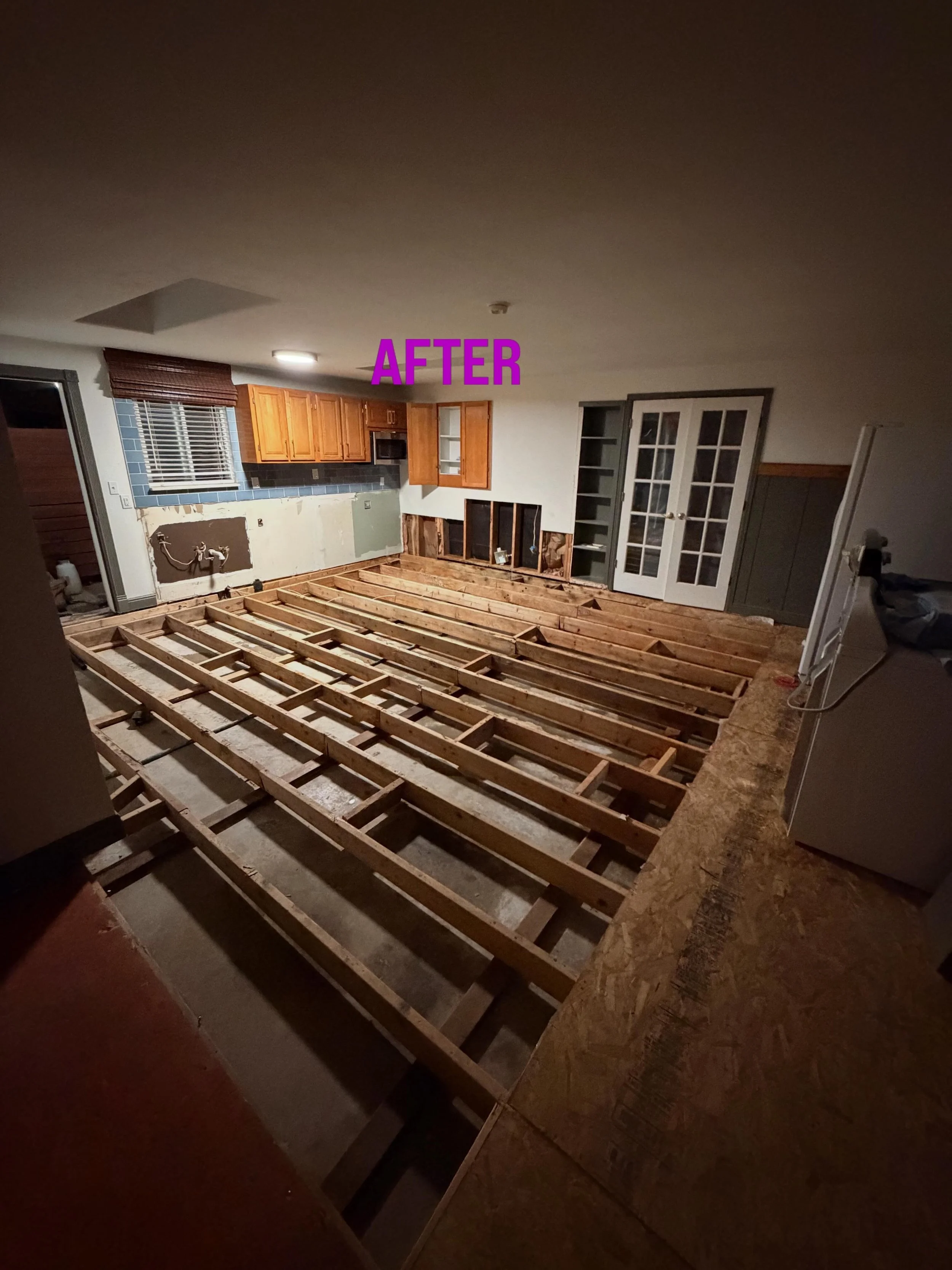 Room under renovation with exposed wooden floor framing, kitchen cabinets, and French doors.