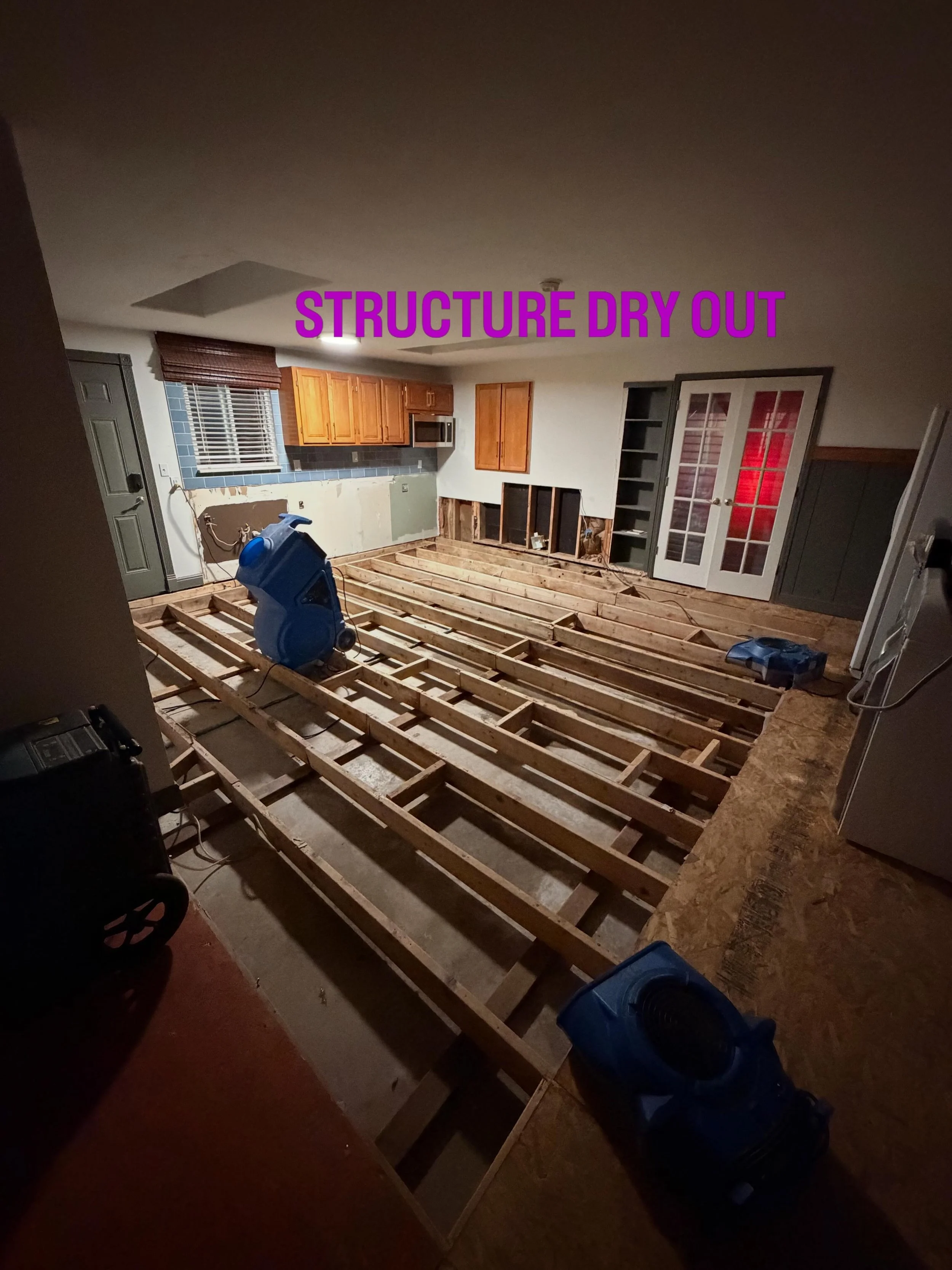 Living room under renovation with exposed wooden floor joists, kitchen cabinets, and removal of flooring or wall panels.