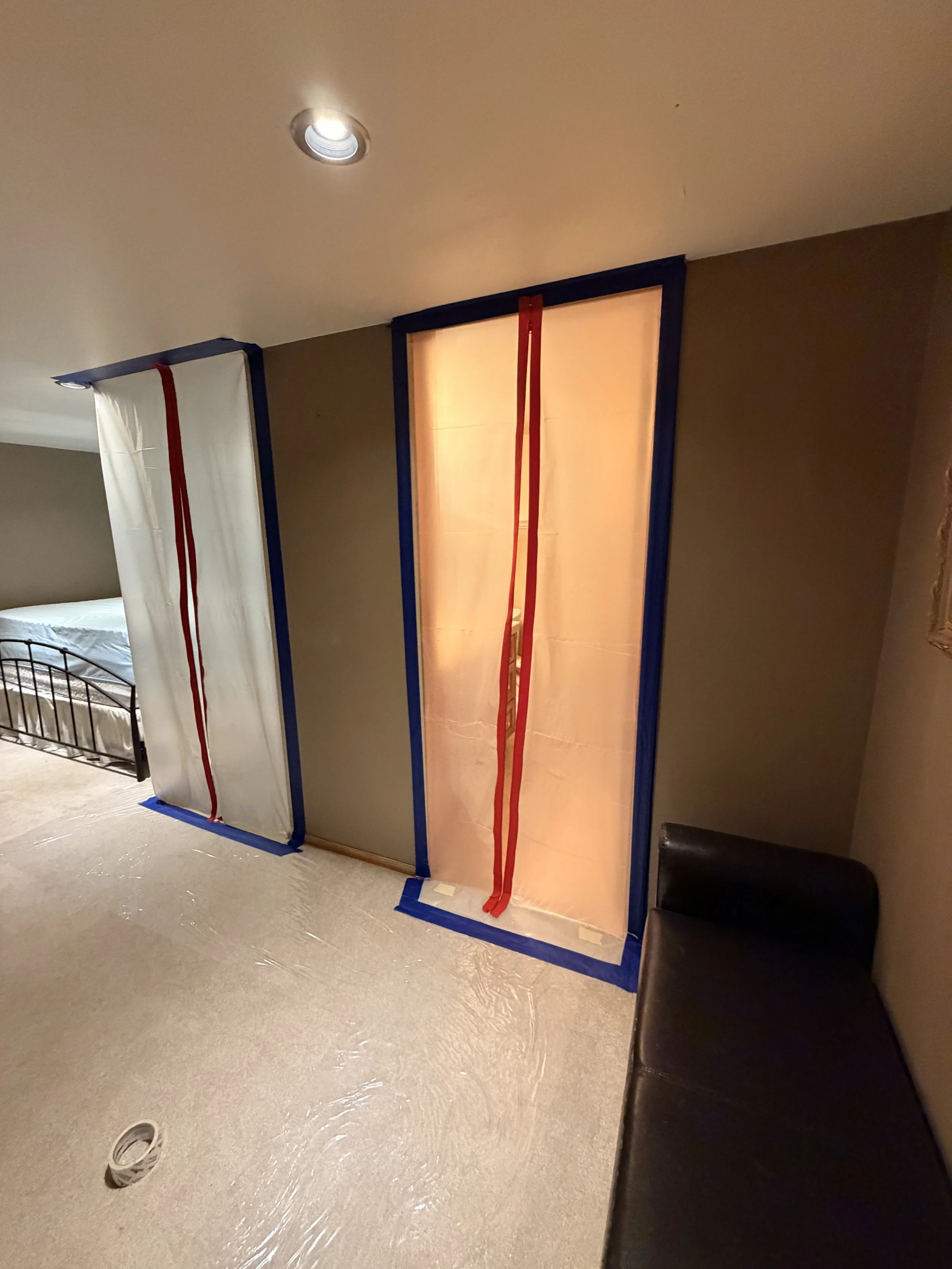 Unfinished room with a partially built partition wall covered in beige paper, framed with blue tape and secured with red straps. To the left, there is a bed with a white sheet and a black metal frame. The floor has plastic sheeting and a circular floor drain.
