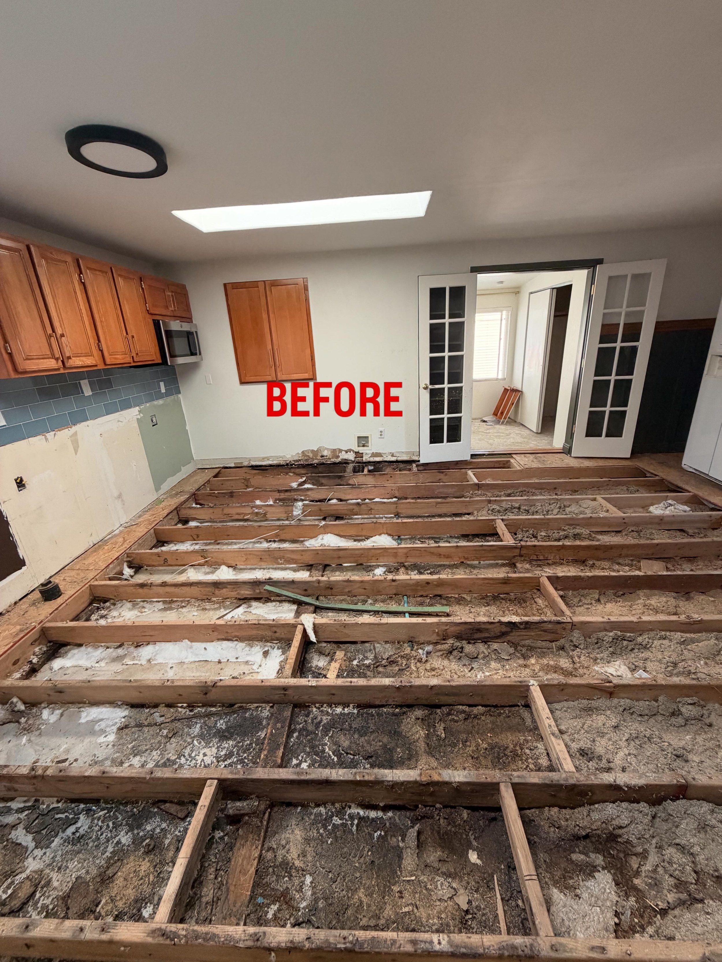 Room with flooring removed, exposing wooden floor joists, bathroom door open to a bright room with window, kitchen cabinets and microwave on wall, marked 'BEFORE' in red.