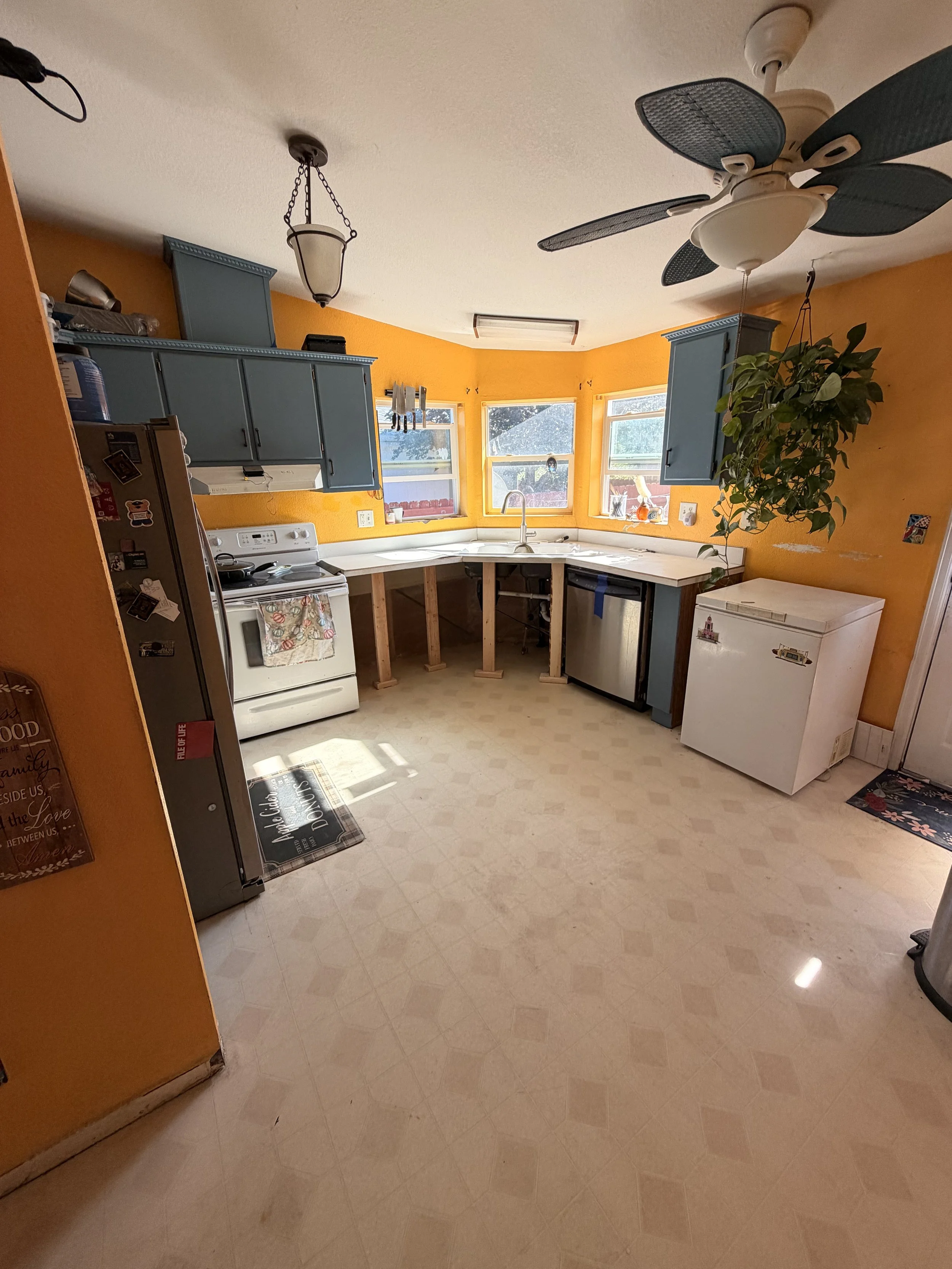 A kitchen with yellow walls, blue cabinets, a white stove with a floral towel, a small white freezer, and a ceiling fan. There are three windows and a hanging plant.