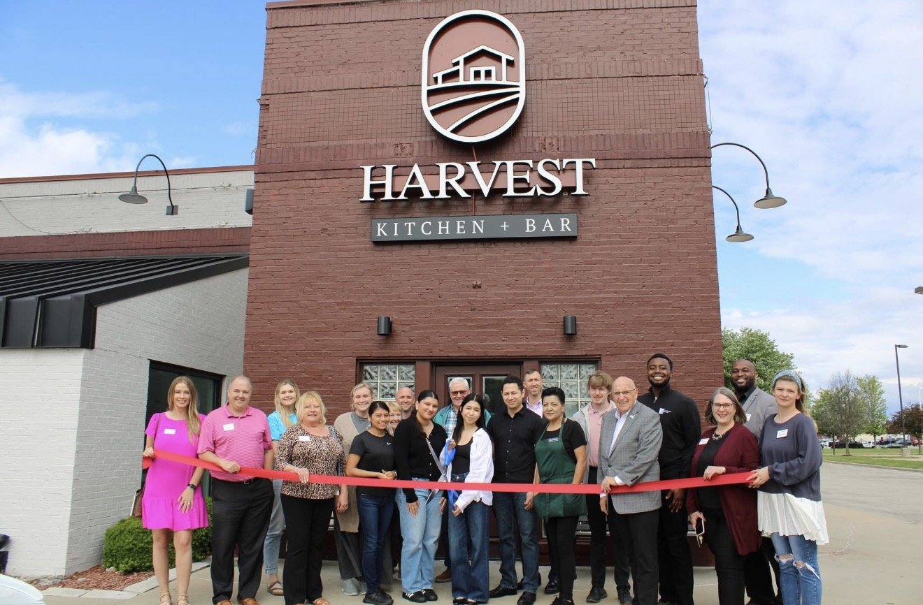 A group of people standing in front of the Harvest Kitchen + Bar restaurant, celebrating a ribbon-cutting event.