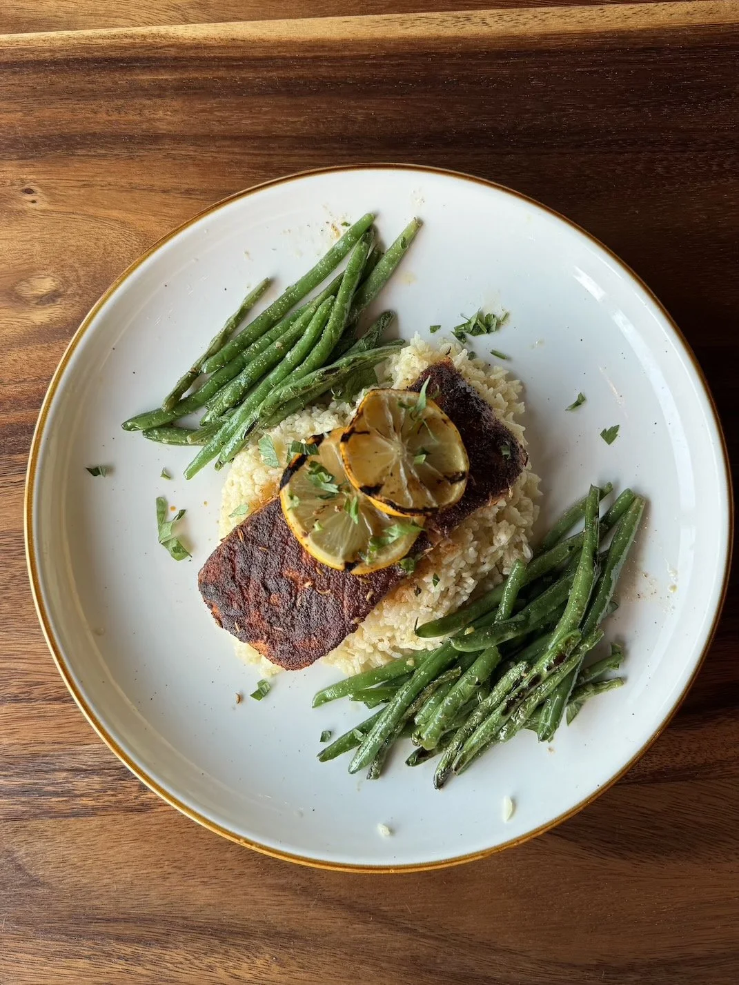 A plate of blackened salmon on a bed of rice with green beans and lemon slices garnished with herbs.