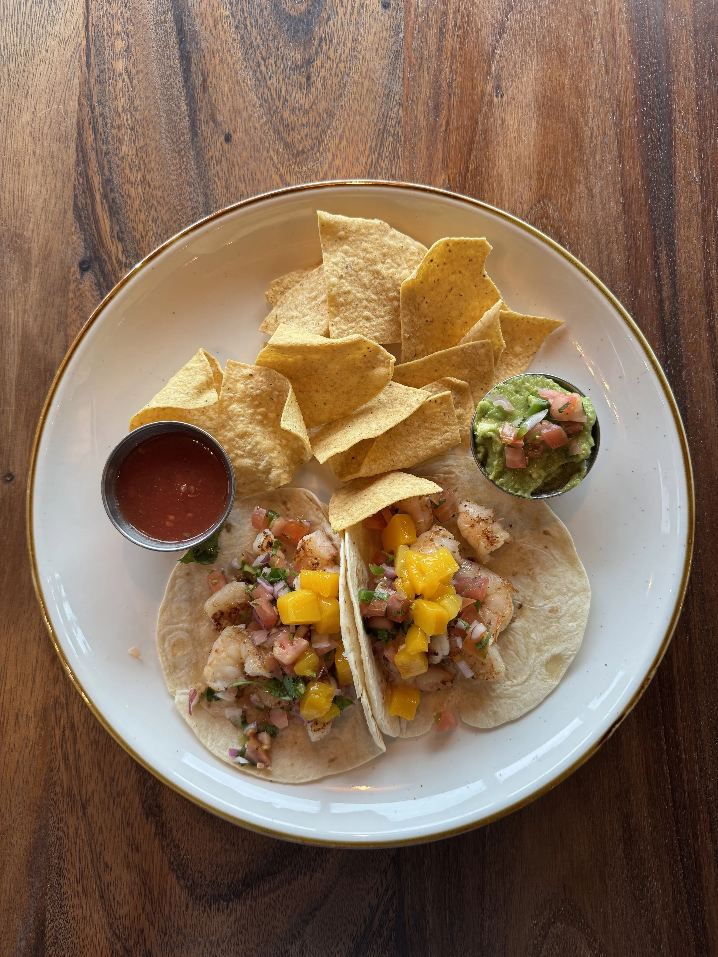 A white plate with two tacos filled with shrimp, diced mango, and chopped onions, accompanied by a side of guacamole topped with chopped tomatoes and onions, and a small bowl of red salsa. The plate is on a wooden table.