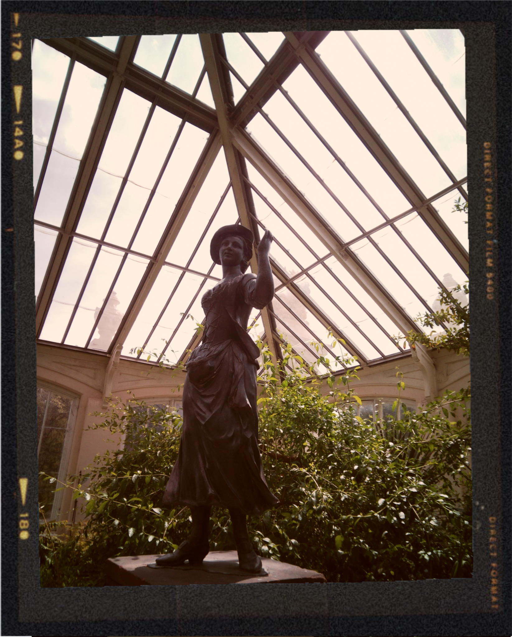 Bronze statue of a woman wearing a hat and boots, standing on a base inside a glass greenhouse with plants around.