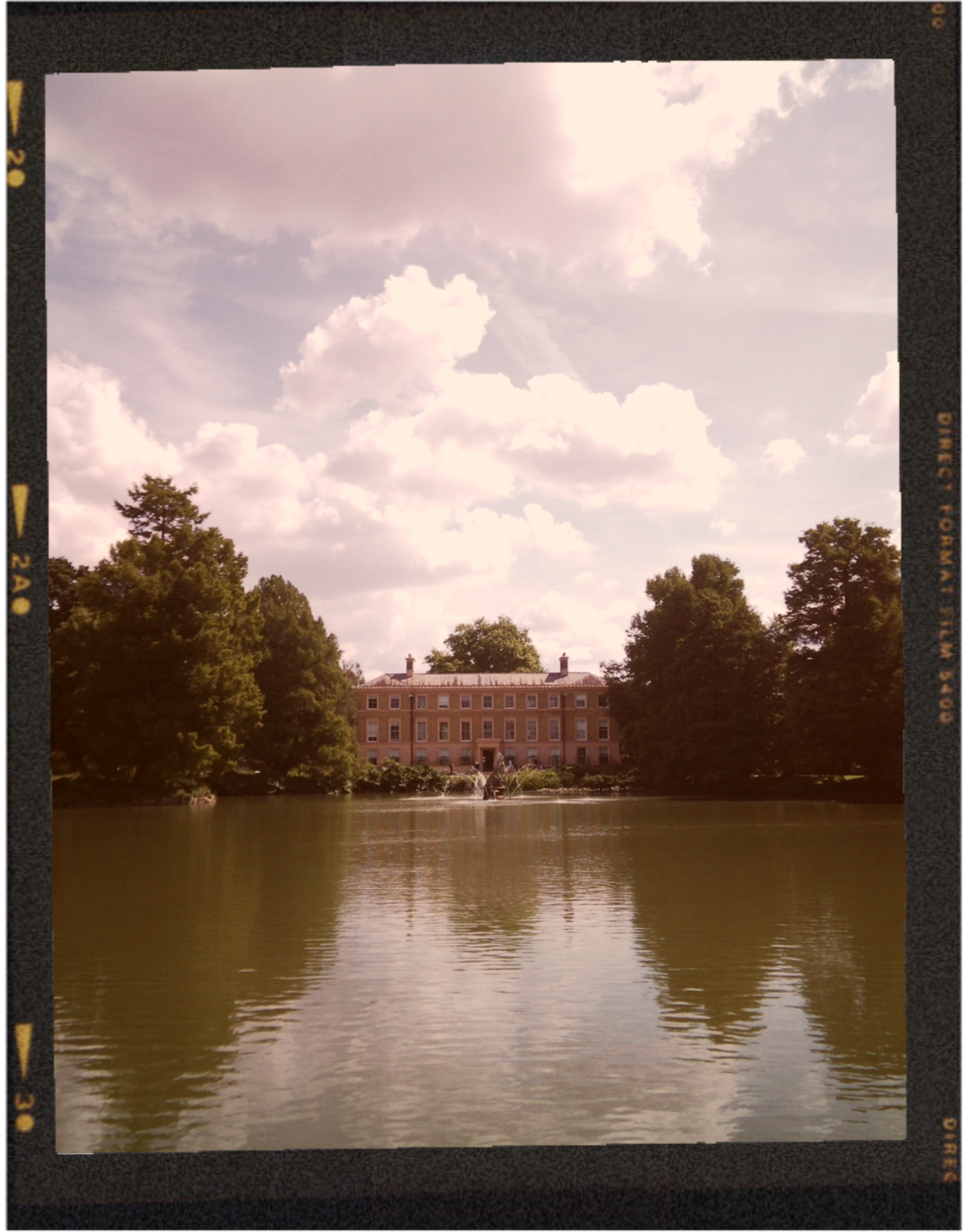 A large historic brick building with multiple windows is reflected in a calm body of water. The scene is surrounded by trees, with a fountain in front of the building, under a partly cloudy sky.