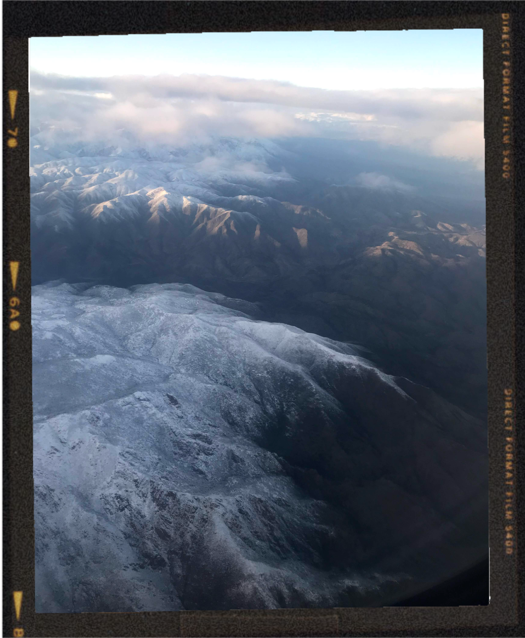 A photo taken from an airplane window showing snow-covered mountains and clouds in the sky.
