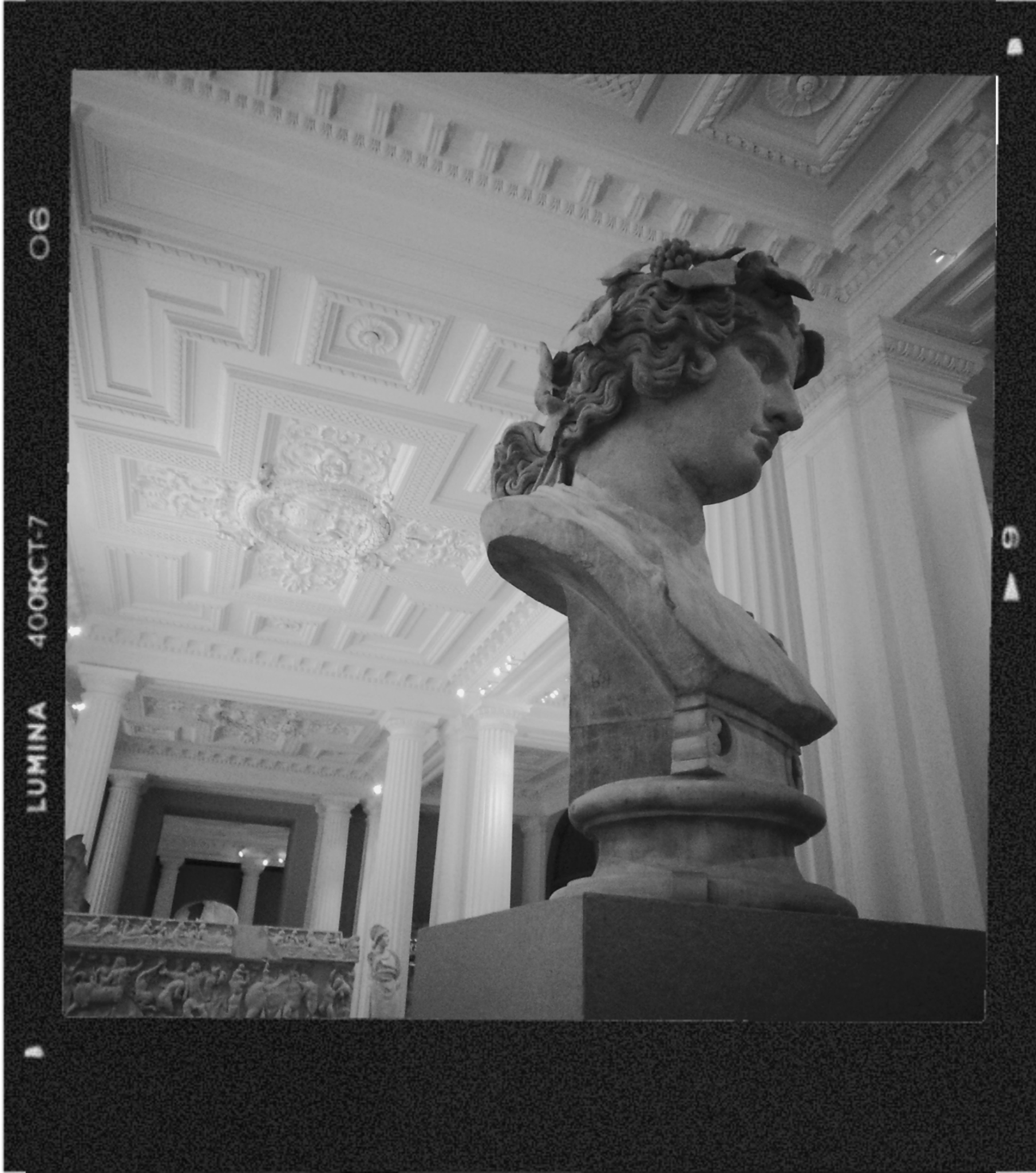 Black and white photograph of a classical sculpture bust of a man with curly hair on a pedestal, inside an ornate hall with decorative ceiling panels and columns.