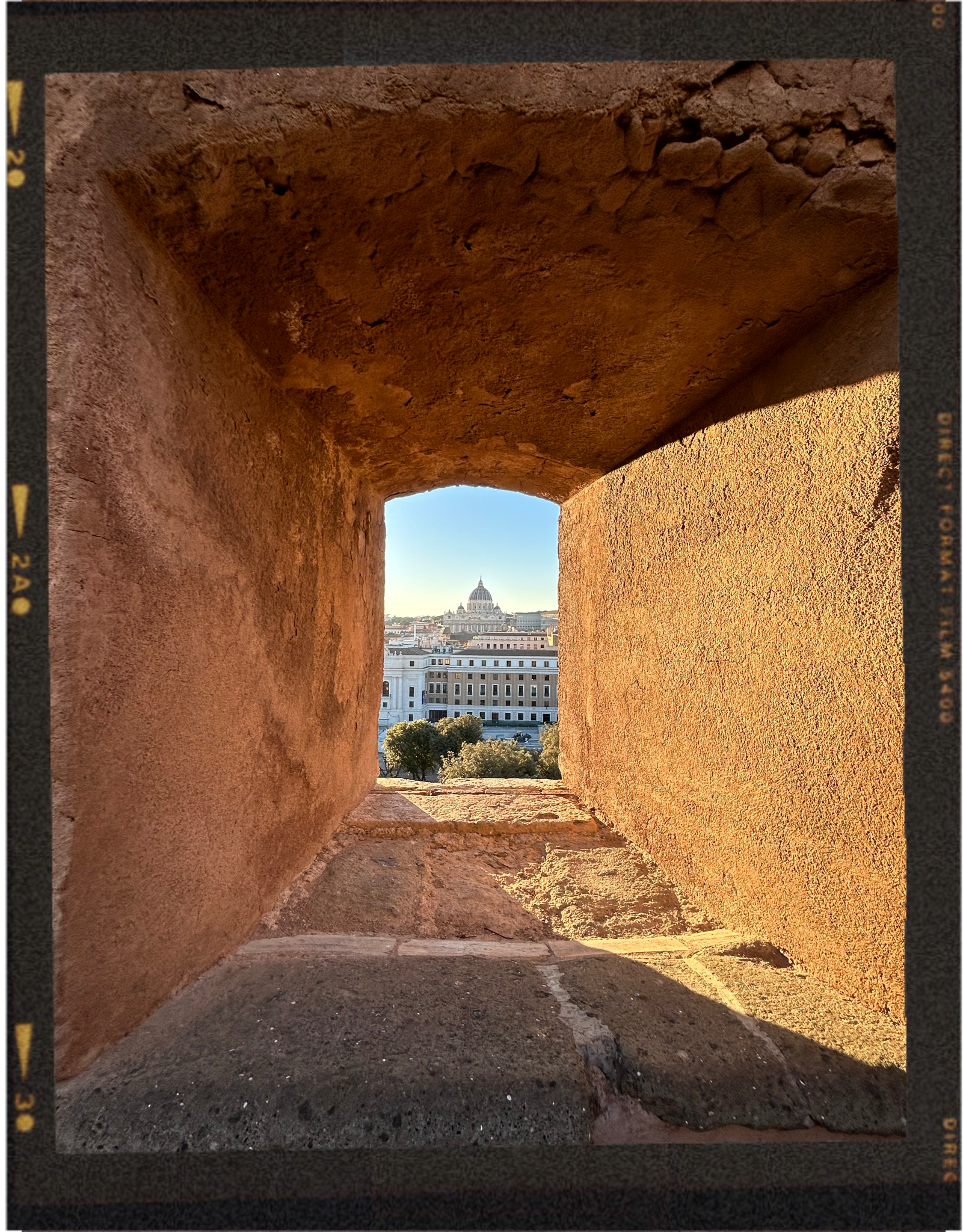 View through a small, rectangular stone window opening in an ancient brick wall, showing a cityscape with a large domed building and trees in the background.