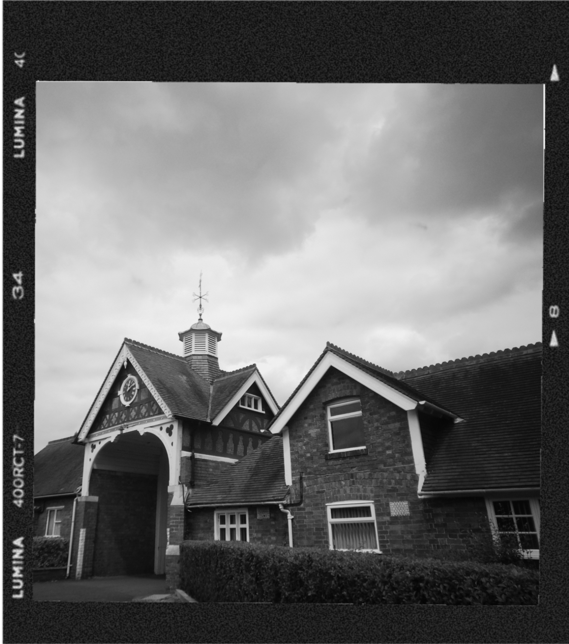 Black and white photo of a large house with a tower and clock, surrounded by a hedged yard and cloudy sky