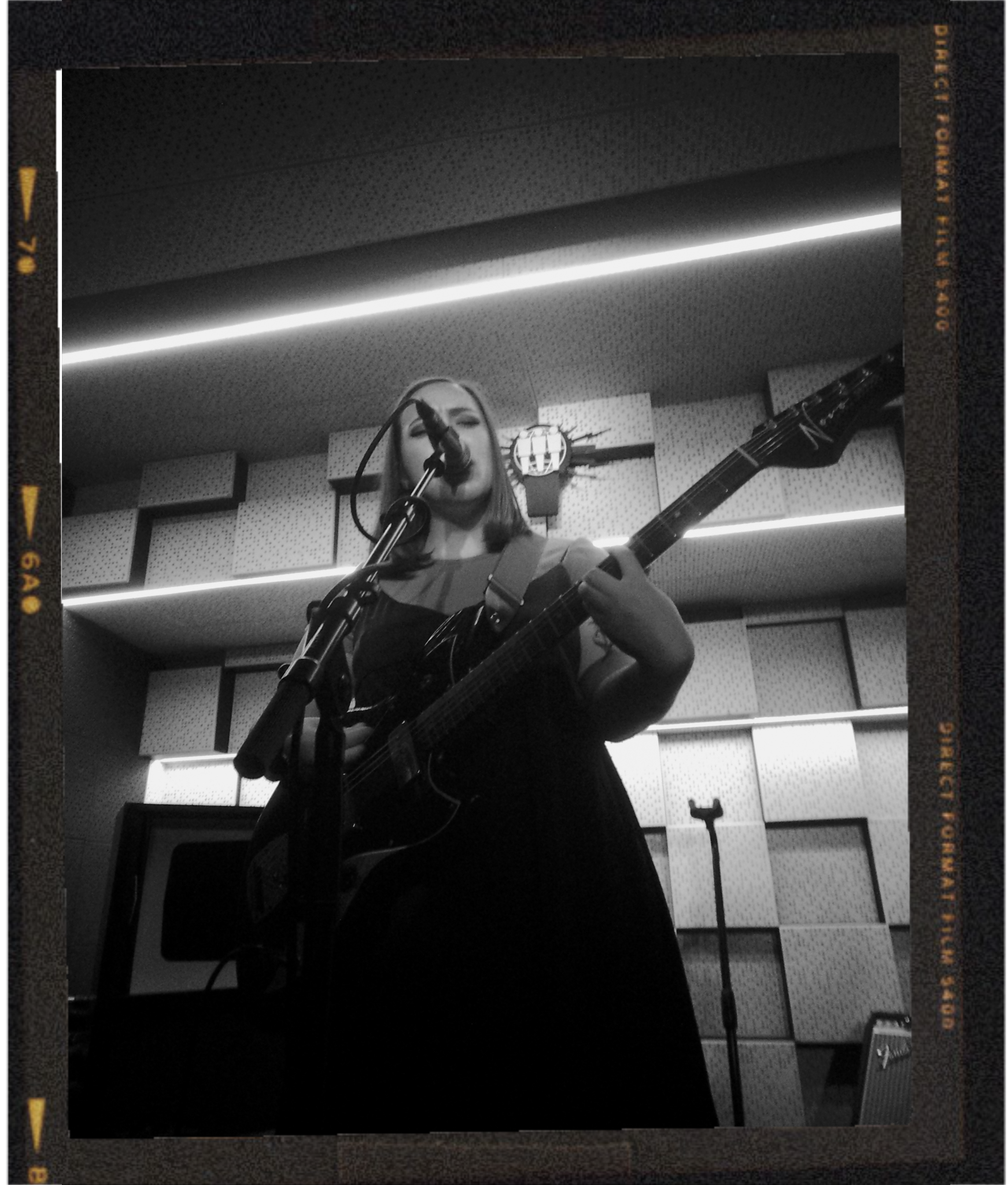 A woman singing into a microphone while playing an acoustic guitar in a recording studio. The studio has acoustic foam panels on the walls and ceiling, with a neon strip light overhead.