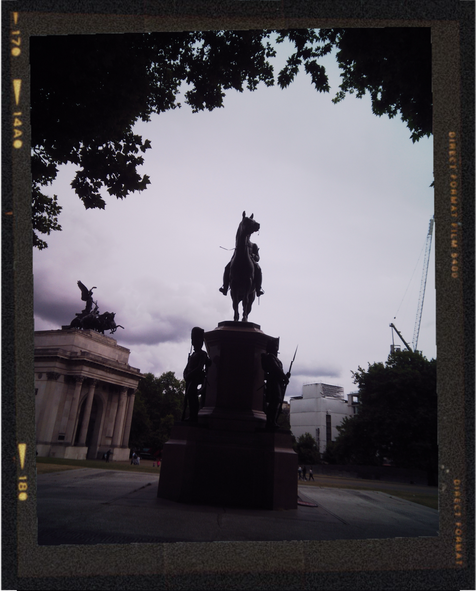 Silhouette of a statue of a person riding a horse, with other statues of soldiers at the base, set against a cloudy sky and framed by trees.