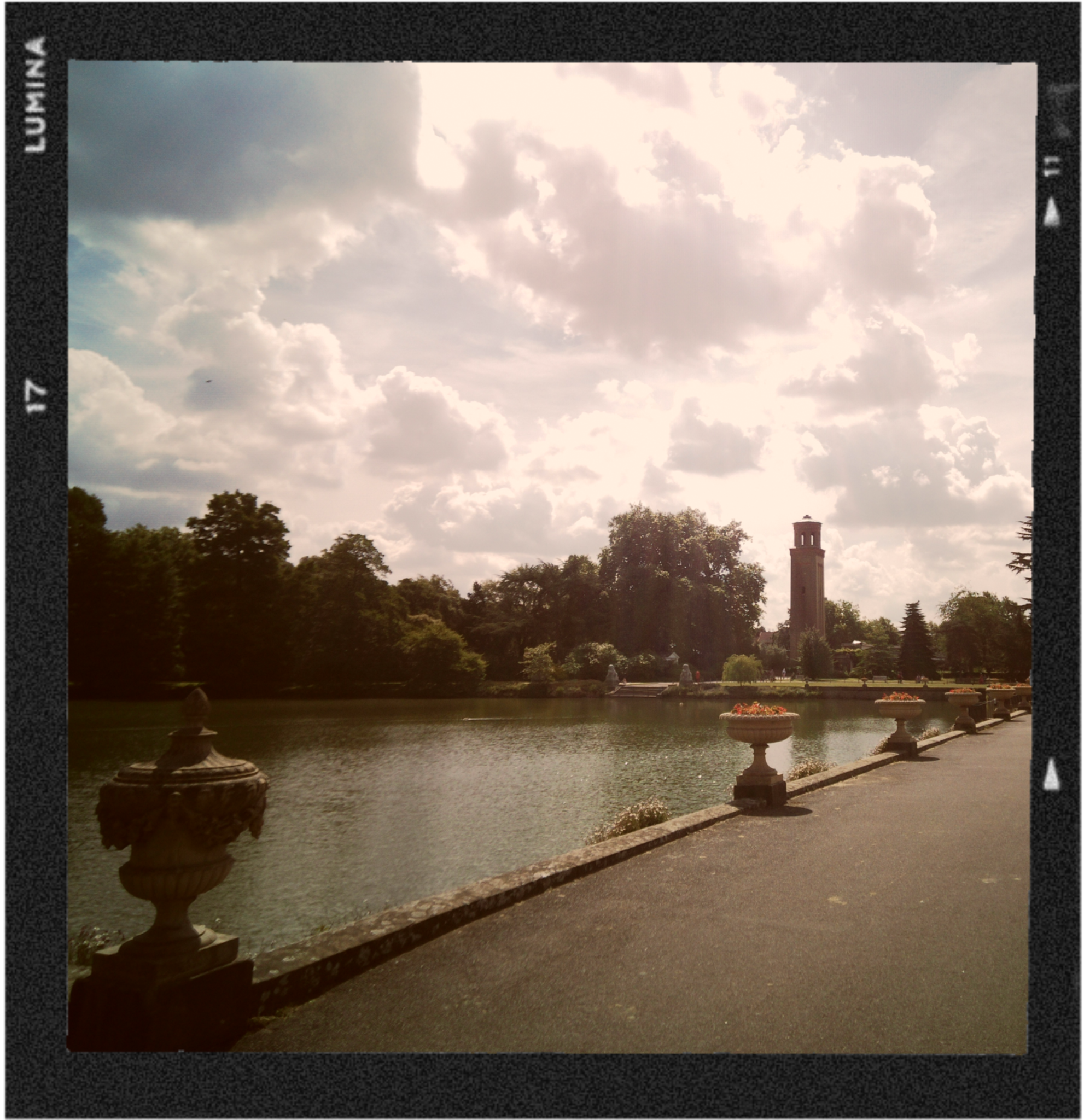 Cloudy sky over a park with a lake, trees, and a tower in the background, viewed from a paved walkway lined with decorative stone planters.
