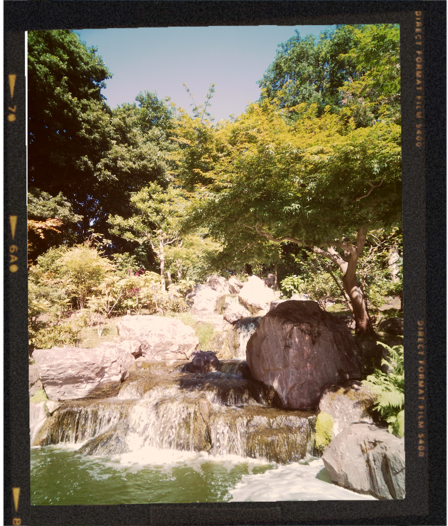 A small waterfall flowing over rocks into a body of water, surrounded by lush green trees on a sunny day.
