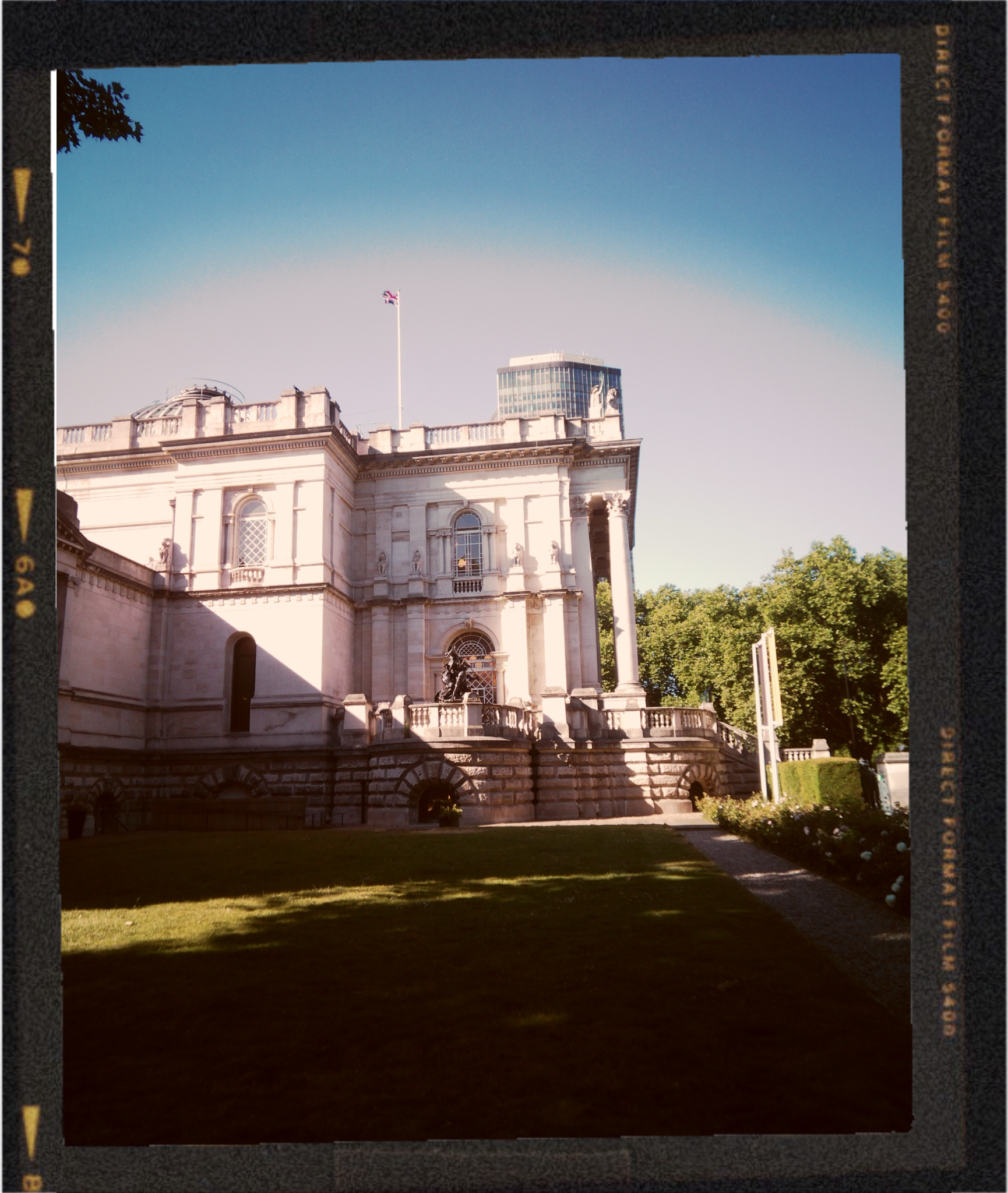 A historic white stone building with classical architectural features such as columns, decorative statues, and ornate windows, with a flagpole flying the British flag on top. In the background, a modern glass skyscraper is visible under a clear blue sky. The scene is illuminated by sunlight, with green trees and a neatly maintained lawn in the foreground.
