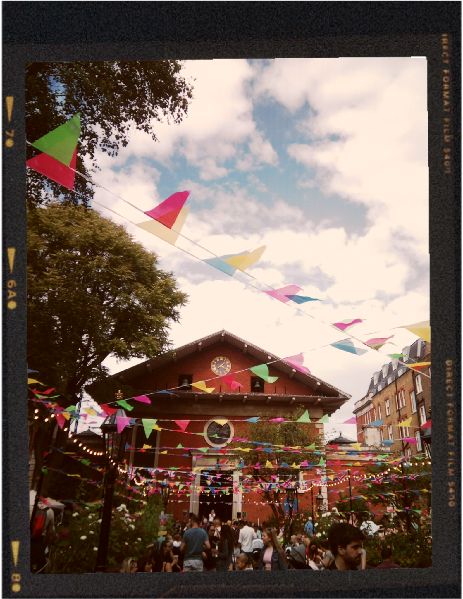 A crowd of people gathered outdoors under a sky with scattered clouds, decorated with colorful bunting flags, in front of a brick building with a clock on its facade.