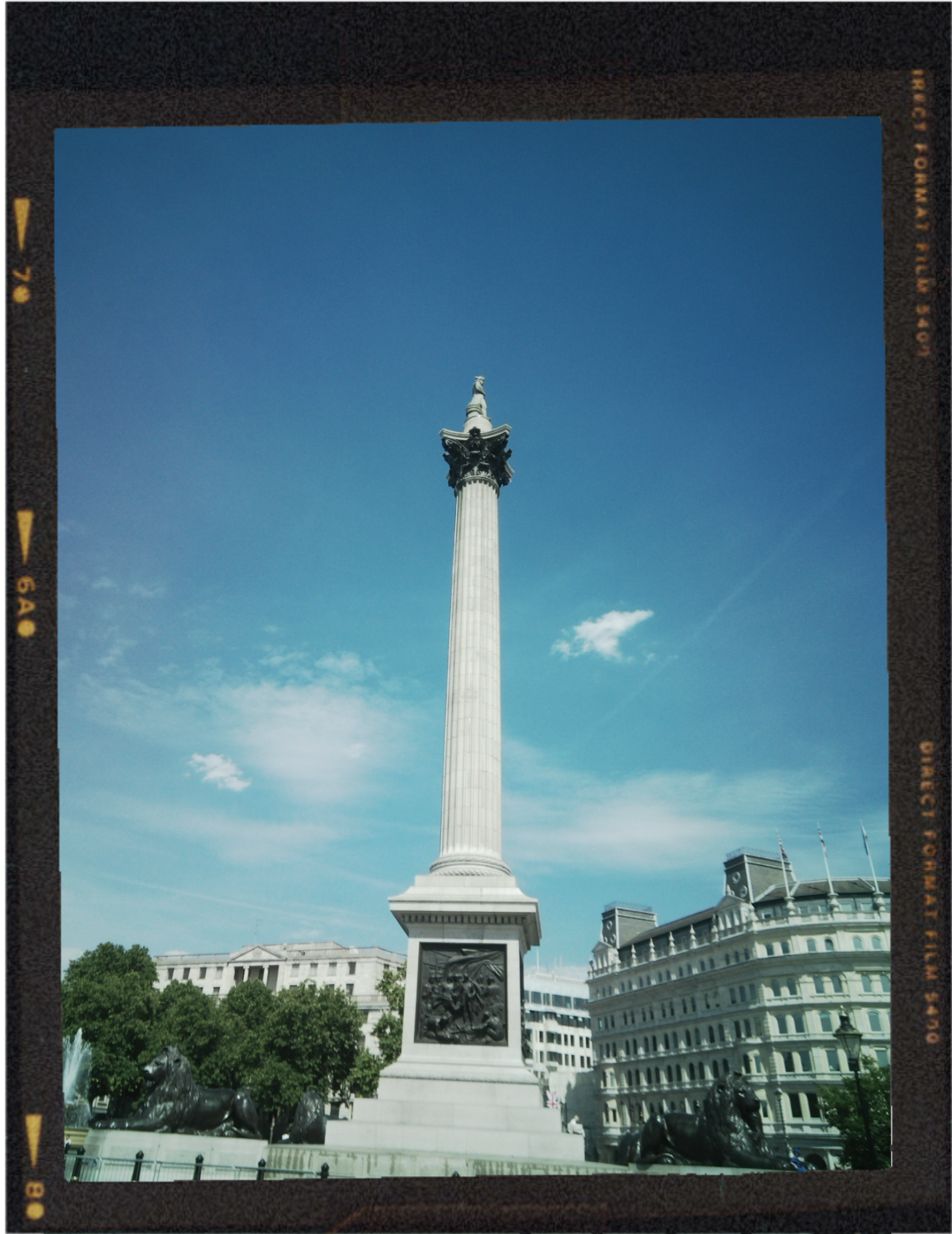 A tall white monument with a statue on top, surrounded by bronze lion statues at its base, located in an urban square with buildings and trees in the background under a blue sky.
