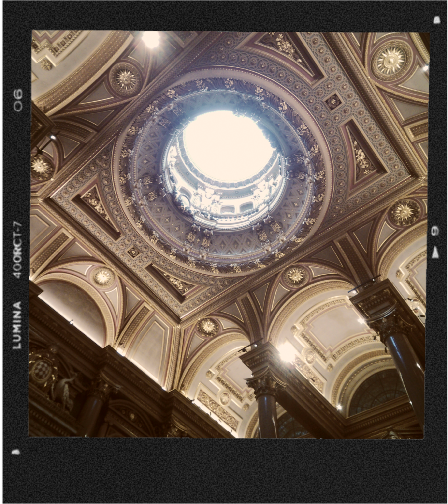 An ornate, decorative ceiling with intricate molding, gold accents, and a circular opening allowing light to shine through, inside a grand building.