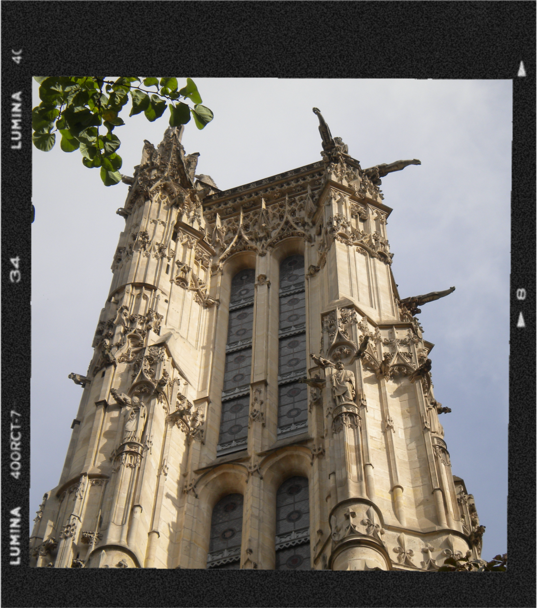 Close-up of the Gothic-style upper section of a church or cathedral with intricate stone carvings and statues, seen from below against a partly cloudy sky with green leaves in the top left corner.