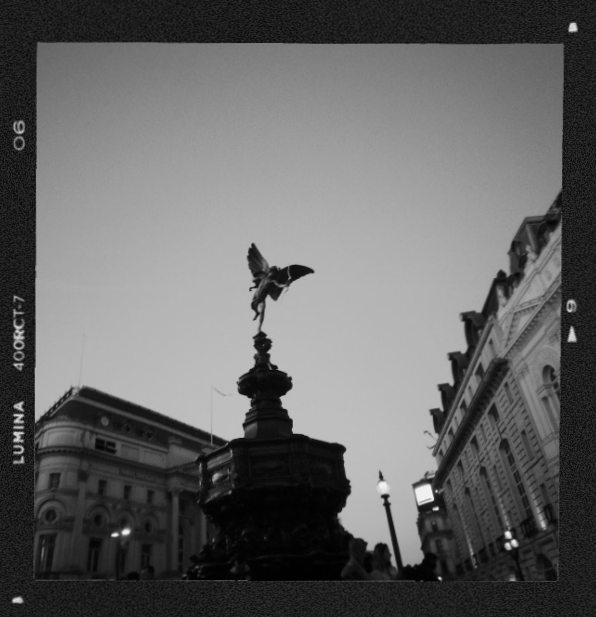 Black and white photo of a city square with a statue of a winged figure on a tall pedestal, surrounded by classical buildings and street lamps.