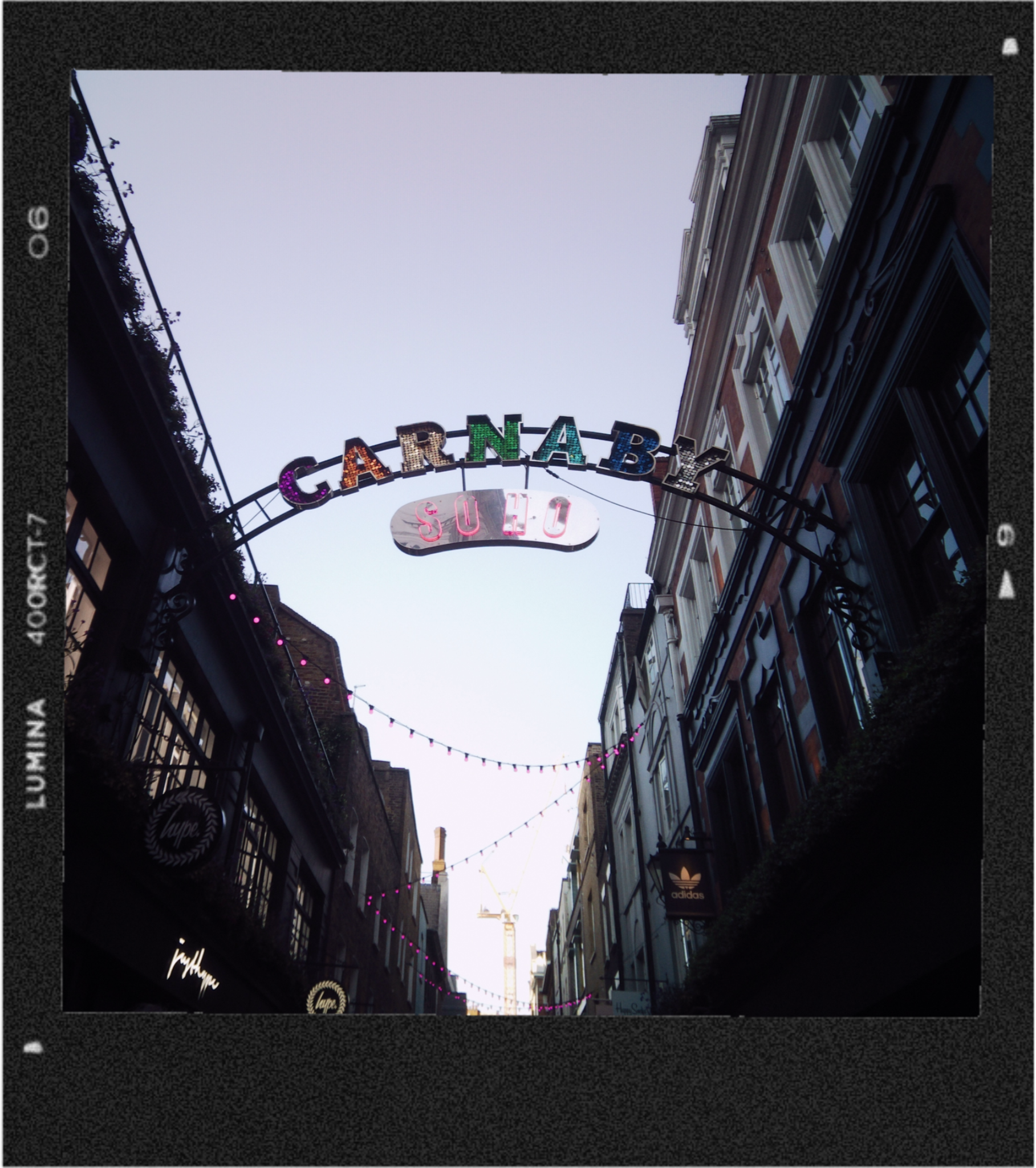 View of a city street with buildings on both sides, colorful illuminated sign that reads 'CARNABY' overhead, and pink string lights. The sky is pale blue, and a construction crane is visible in the distance.