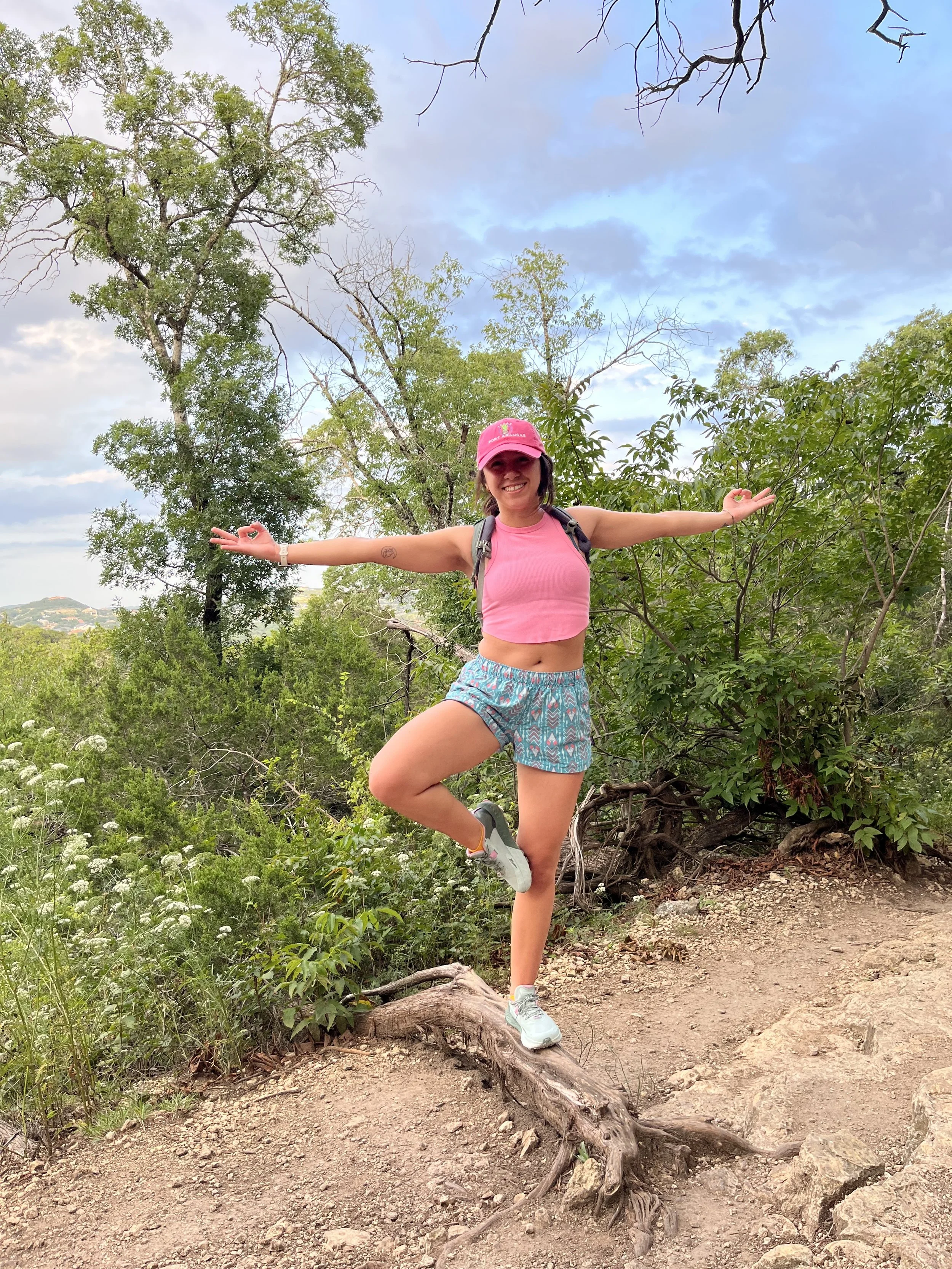A young woman in a pink cap and pink tank top, with patterned shorts, is standing on one leg on a log on a dirt trail, with arms outstretched in a yoga pose or balancing pose, surrounded by green trees and bushes, with a partly cloudy sky in the background.
