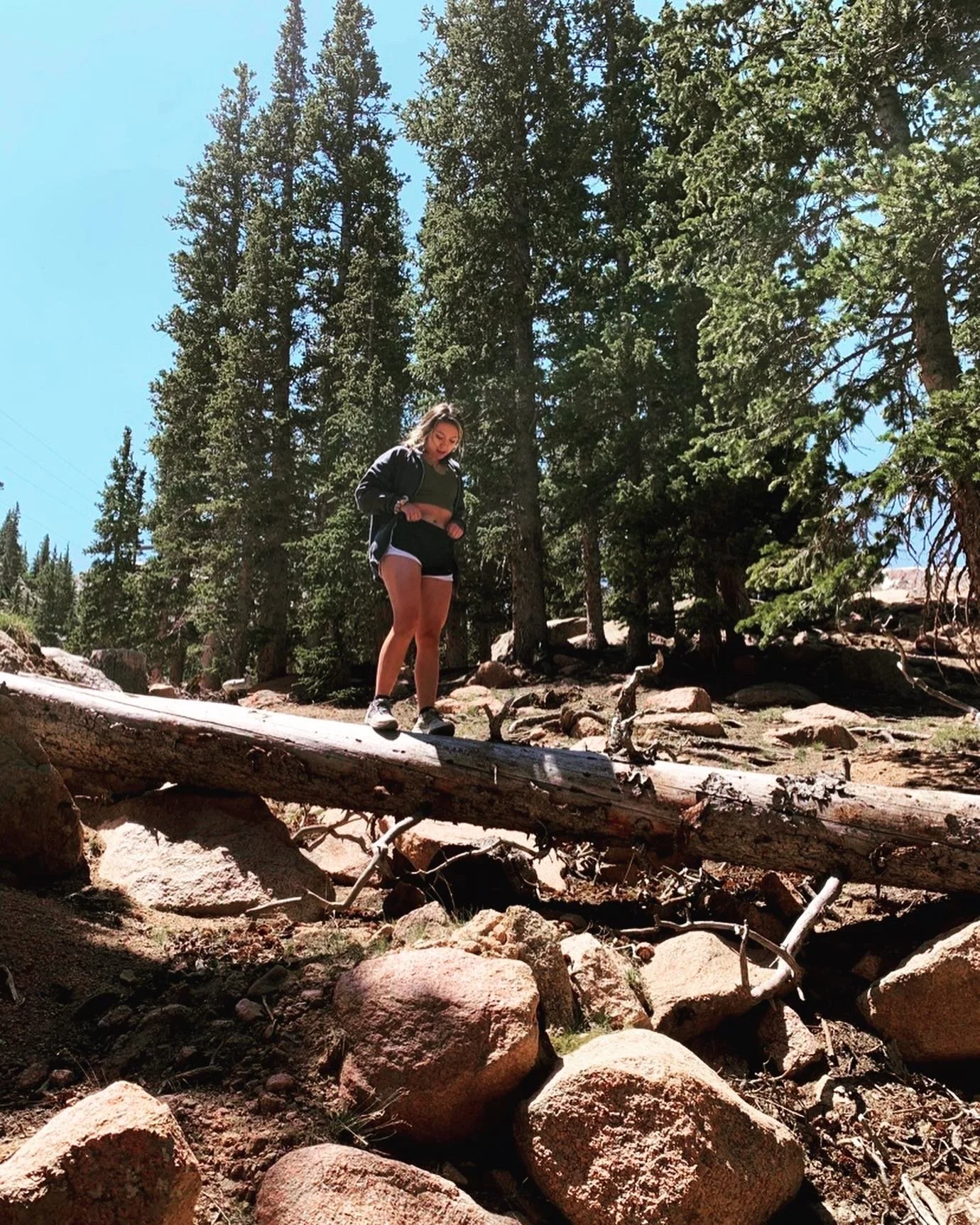 A girl standing on a fallen log in a forest with large rocks and tall pine trees, under a clear blue sky.