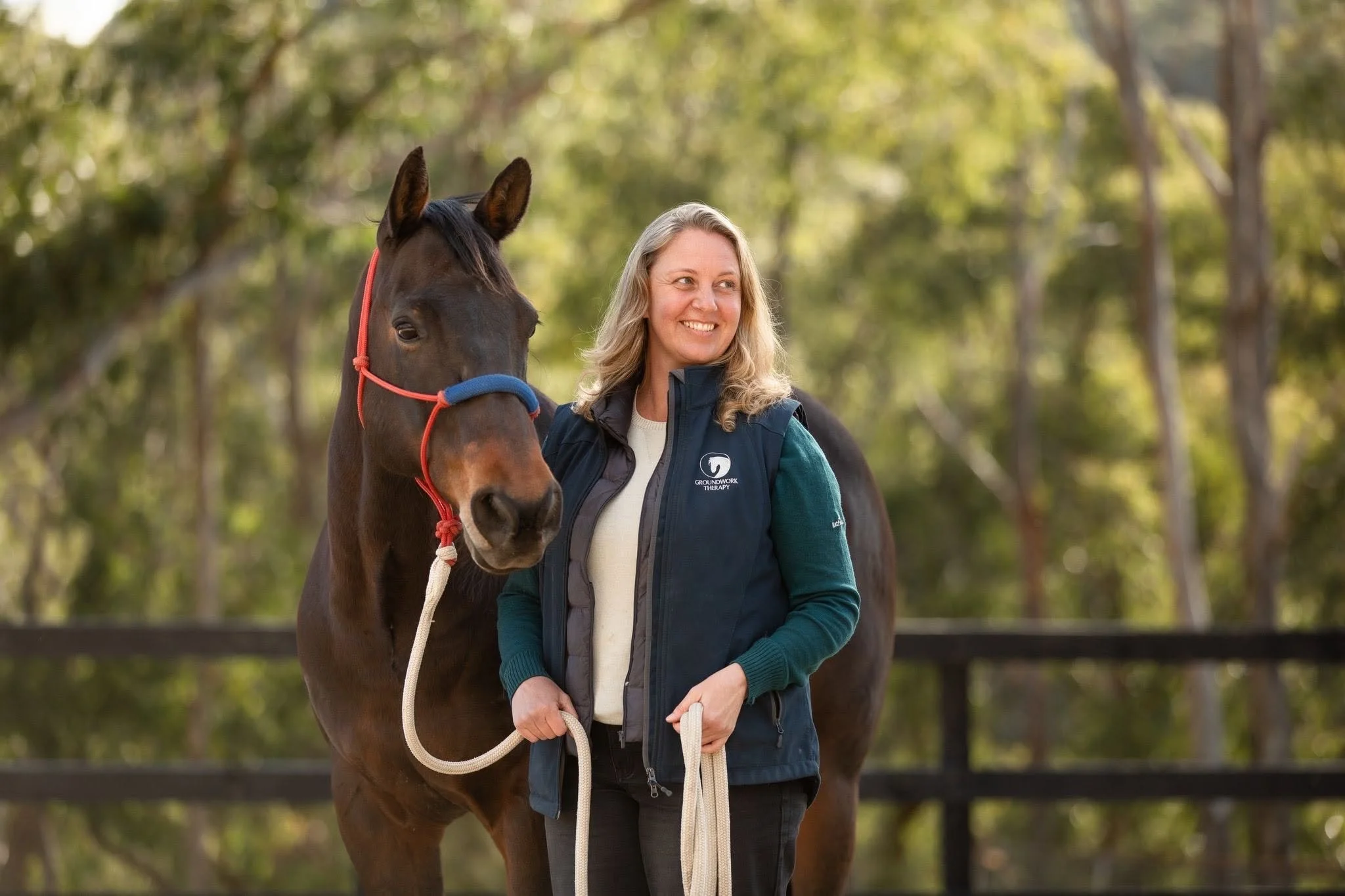 A woman with blonde hair smiling and standing next to a brown horse outdoors with a background of trees and green foliage.
