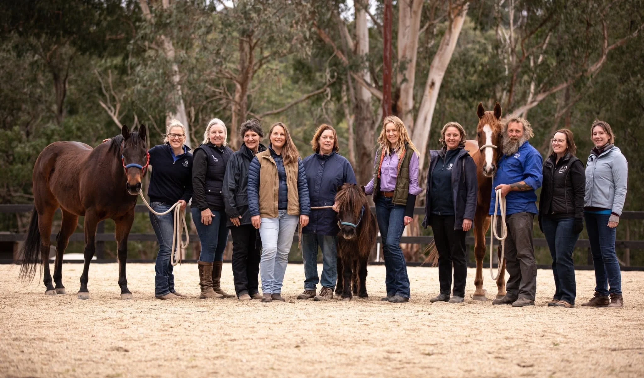 A group of ten people standing with three horses in an outdoor riding arena, with trees in the background.