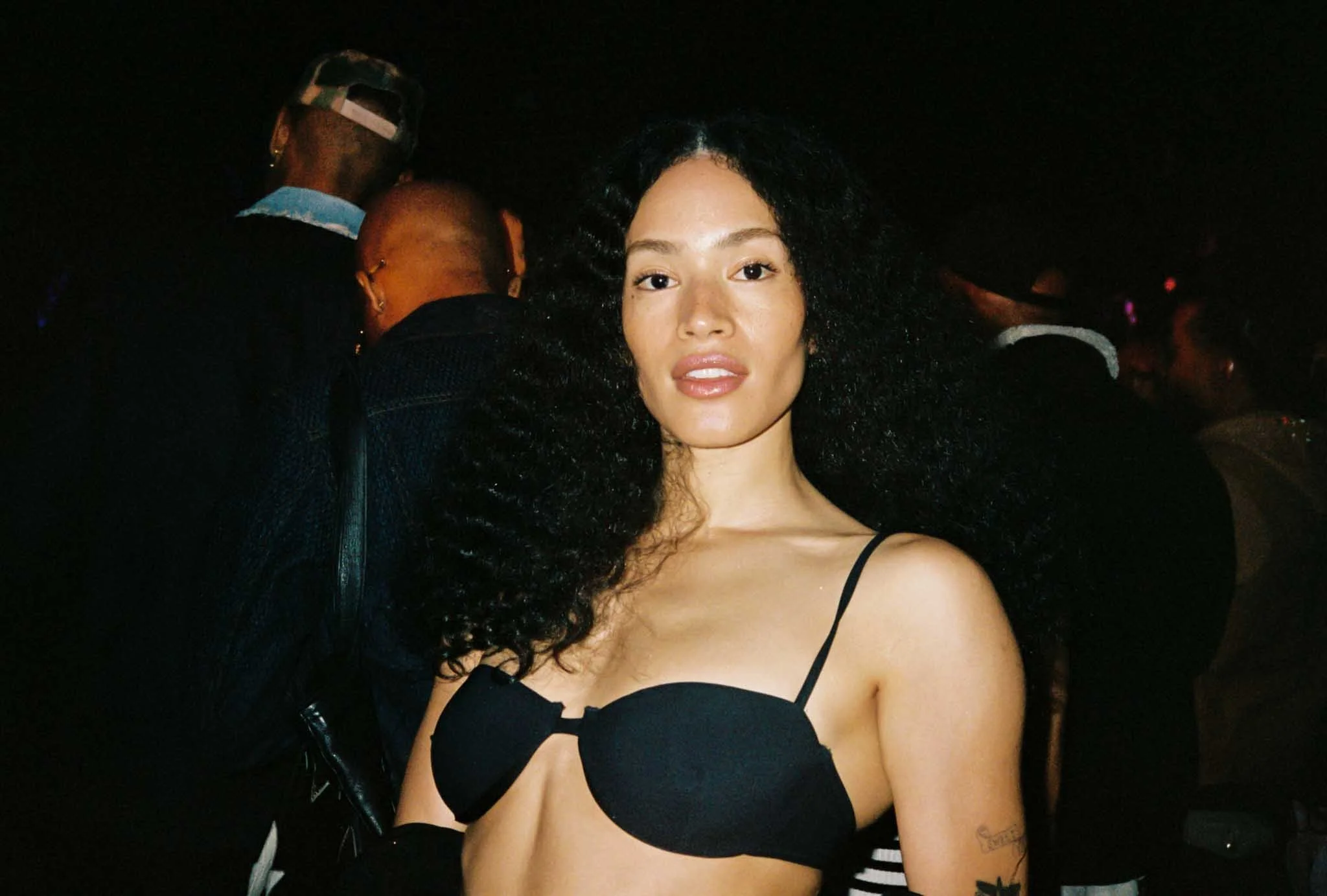 A woman with curly black hair wearing a black spaghetti strap top, standing in a dark environment with other people blurred in the background.