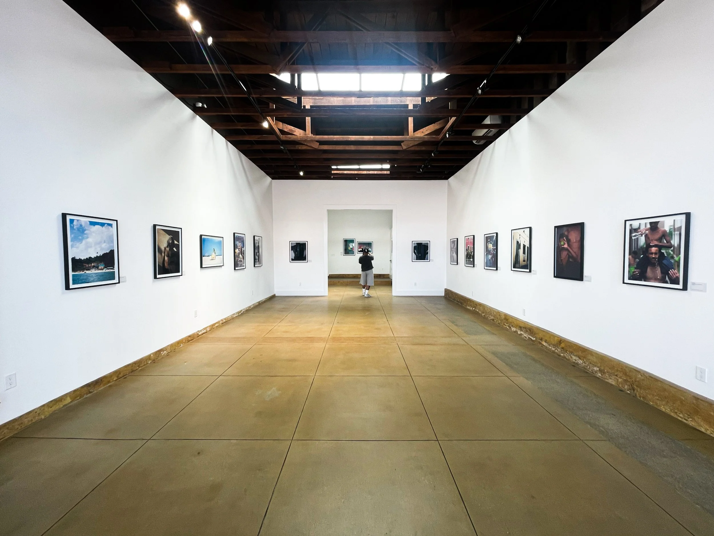 Art gallery with framed photographs on white walls, concrete floor, exposed wooden ceiling, and a person viewing art in the distance.